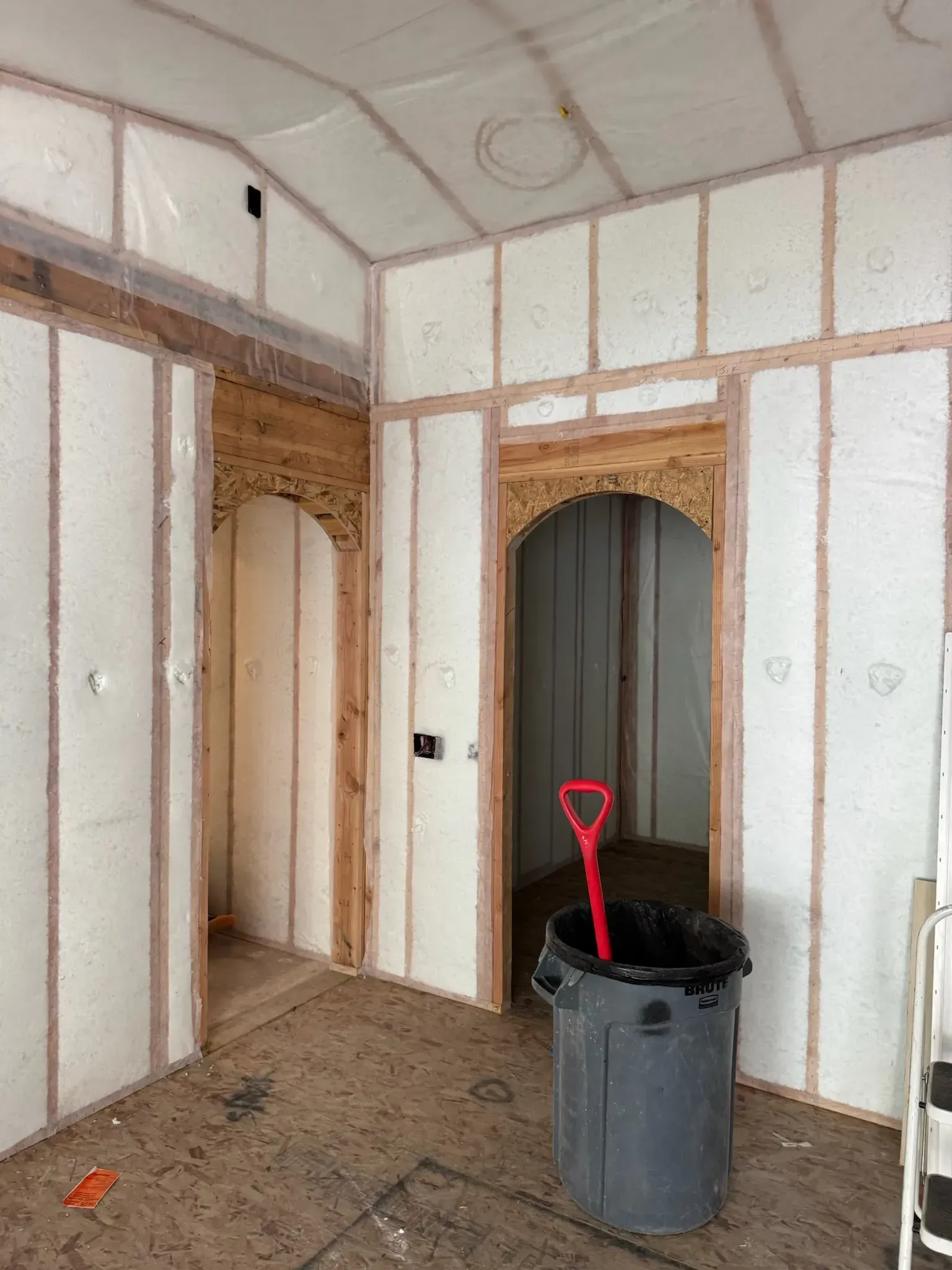 Unfinished room with exposed framing, arched doorway, and a gray bucket with a red shovel in the foreground