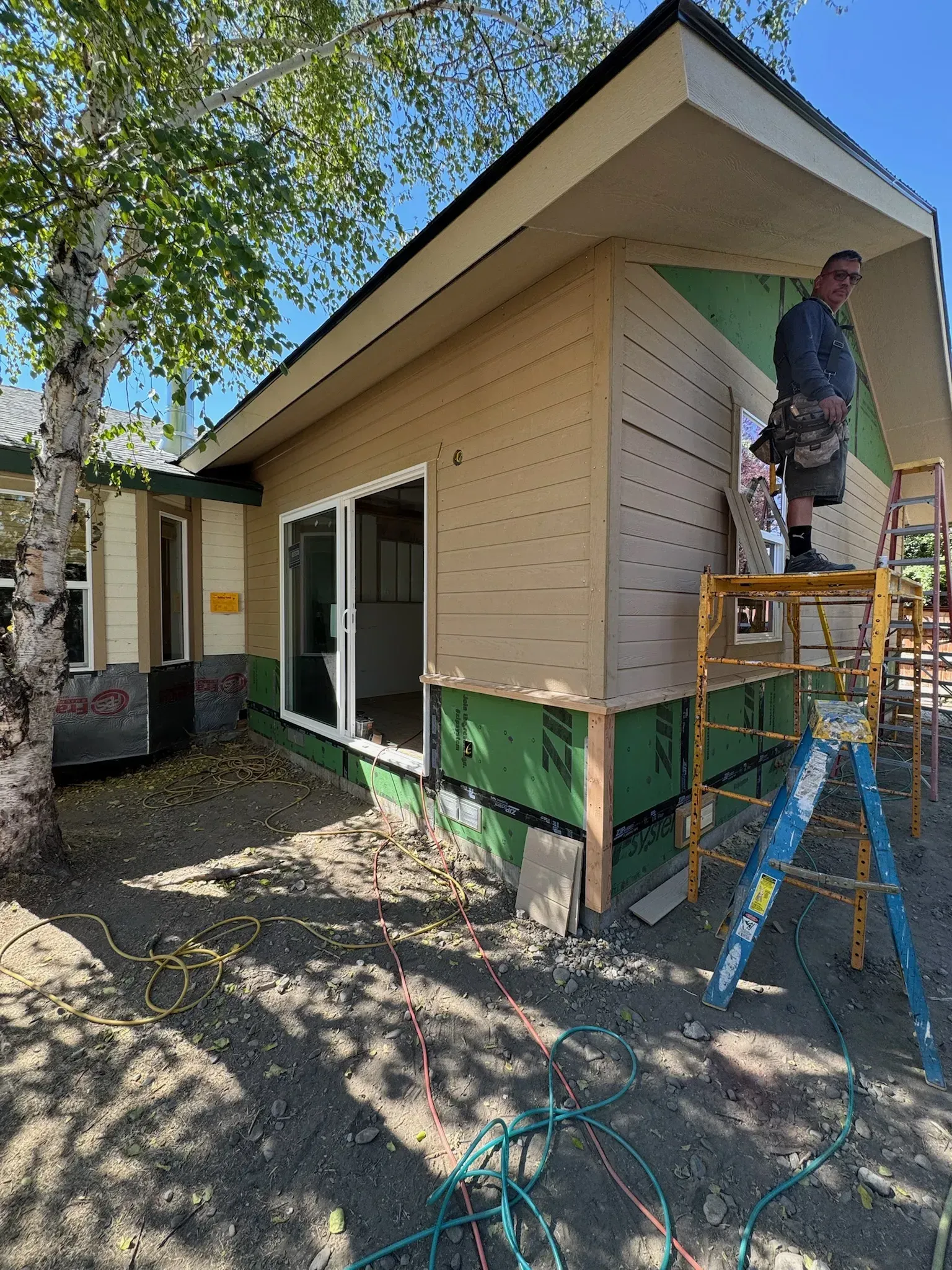 Worker painting exterior of a tan house on a ladder beside a green-trimmed wall
