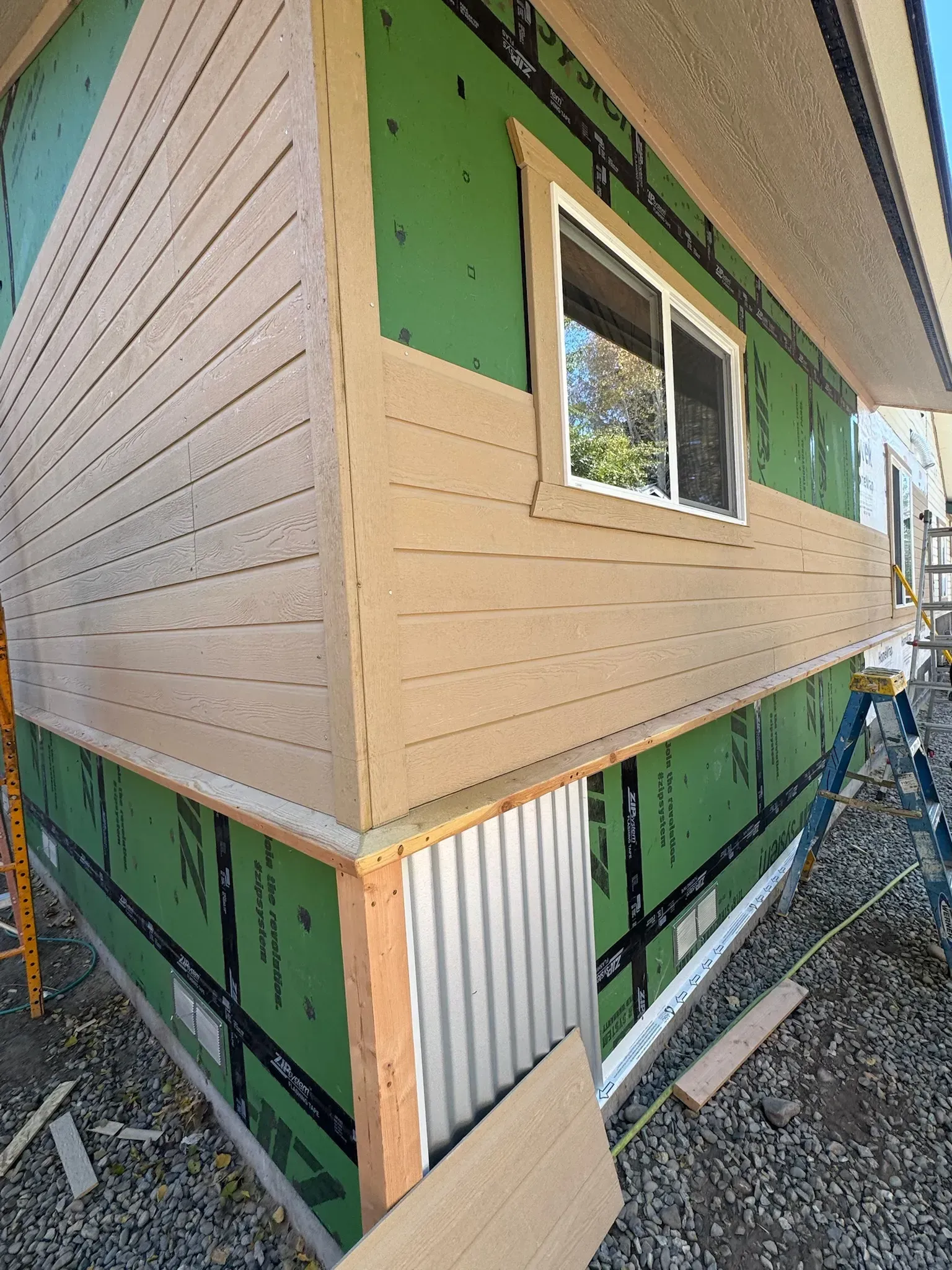Corner of house under construction with tan siding, green sheathing, and a window installed