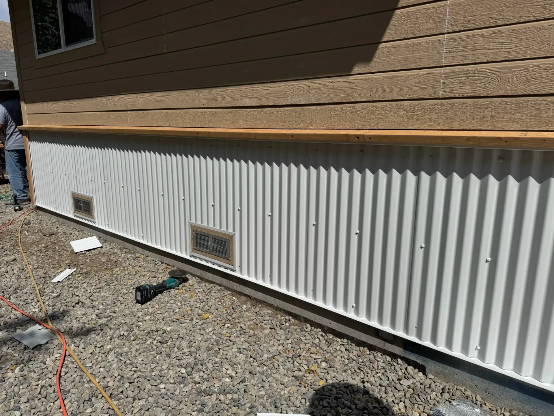 White corrugated metal skirting along a house exterior on gravel, with small vents and a person partly visible at left