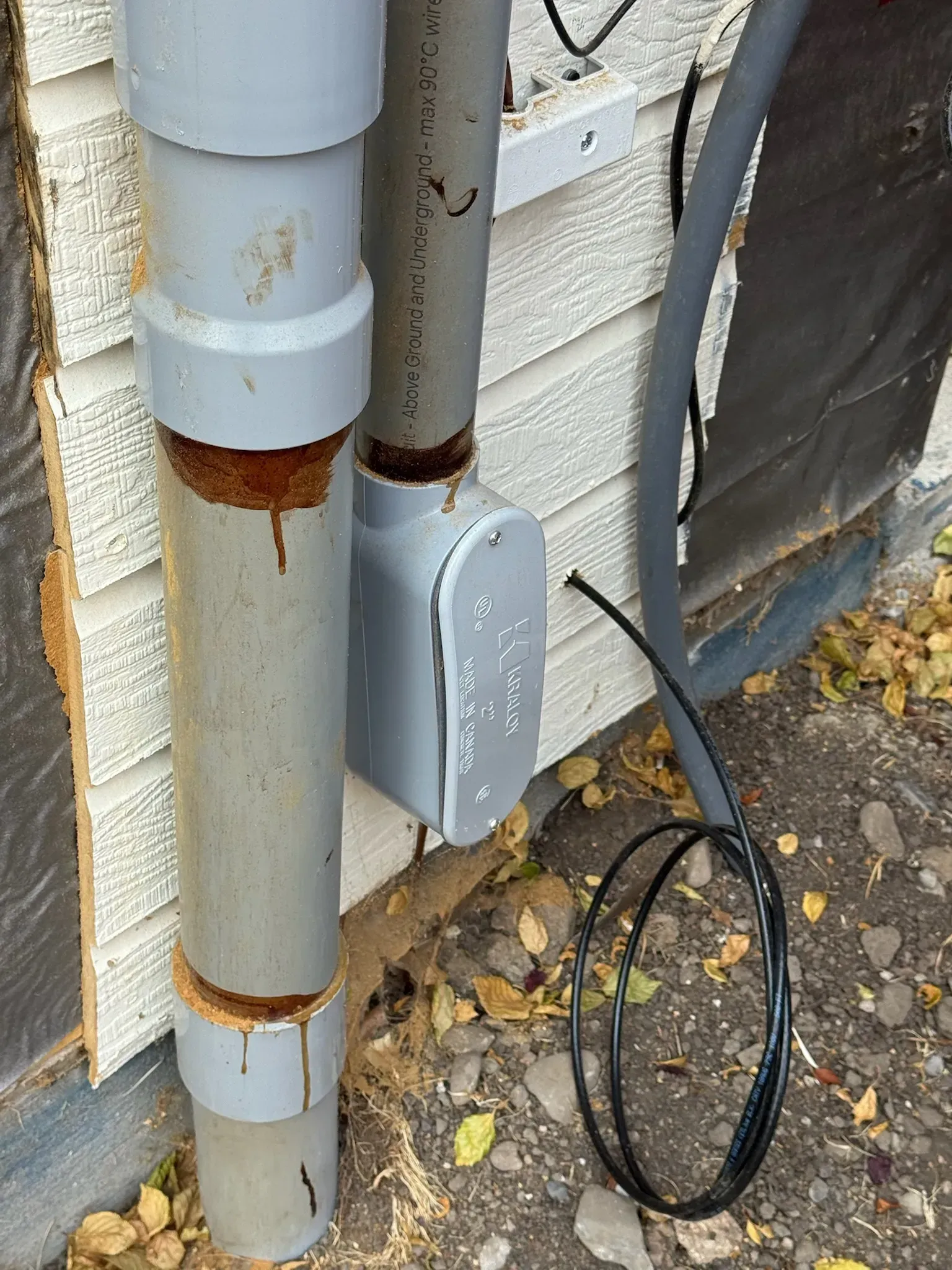 Rusty pipes and gray utility box with a black cable beside a house wall.