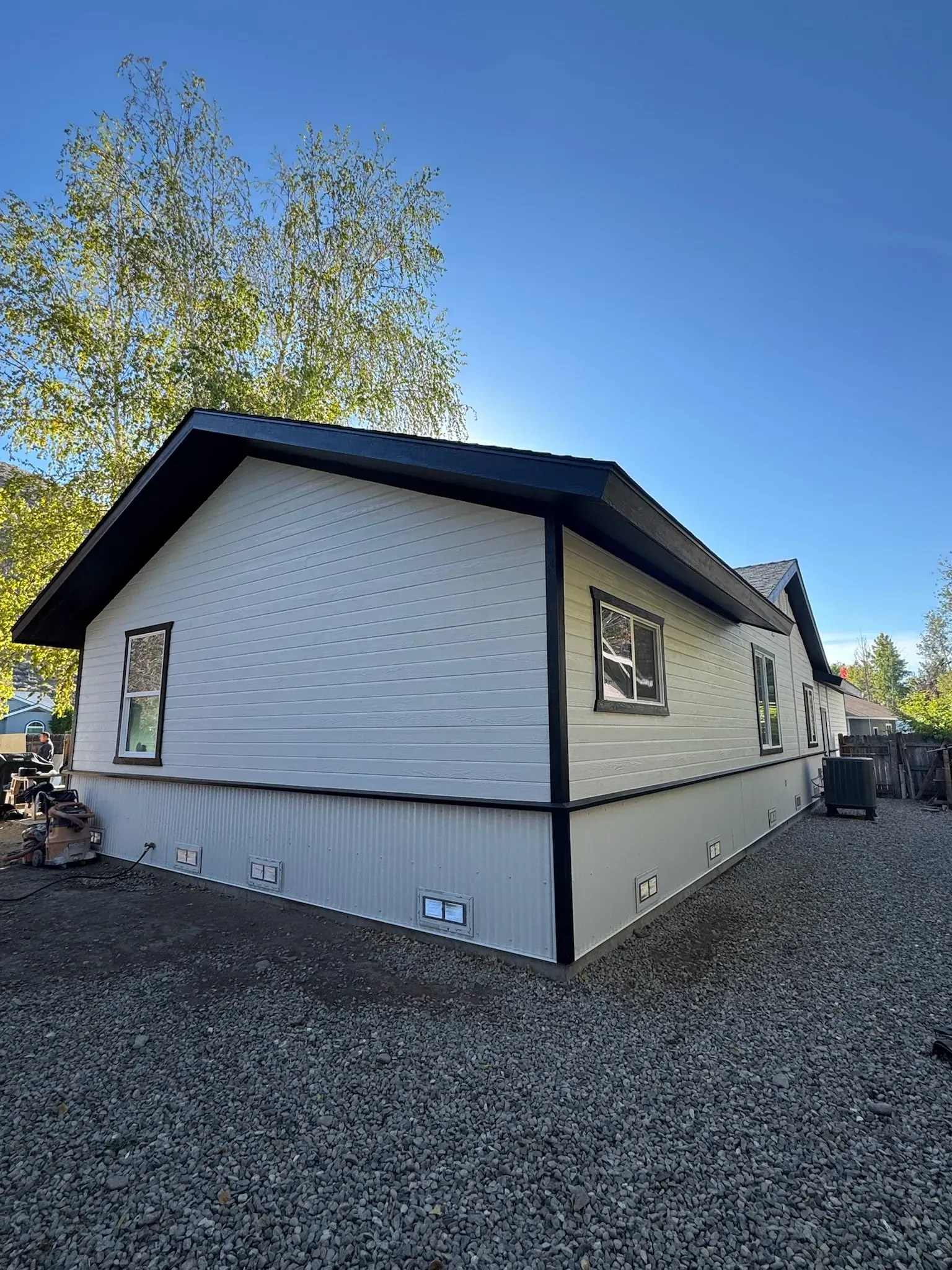 White house with black trim and skirting, seen from the side on a gravel lot under a blue sky.