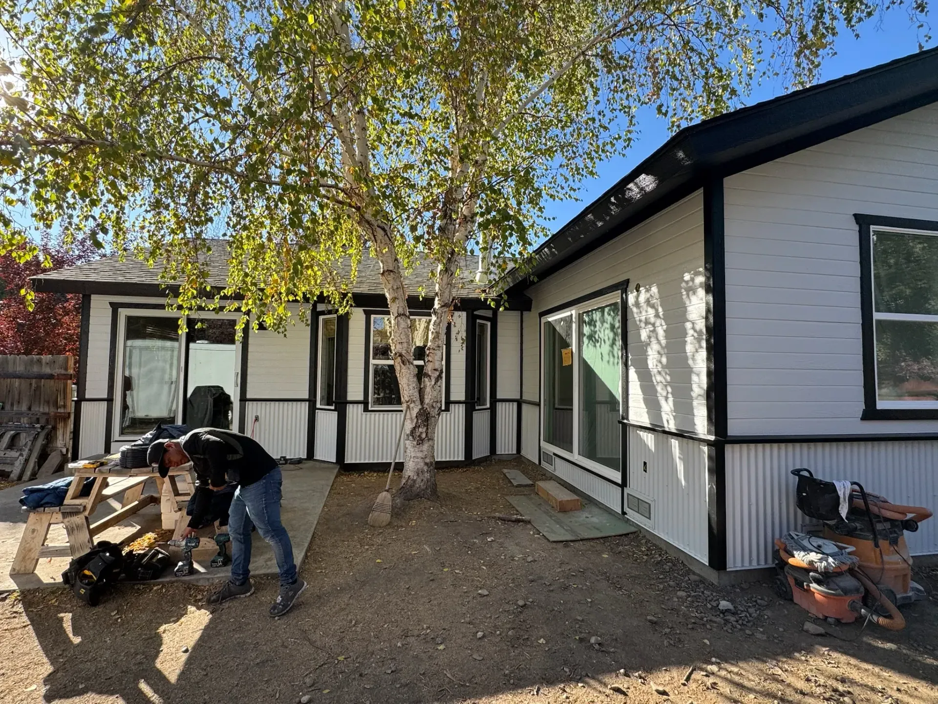 Backyard patio with workers stacking wood and a white house under a leafy tree
