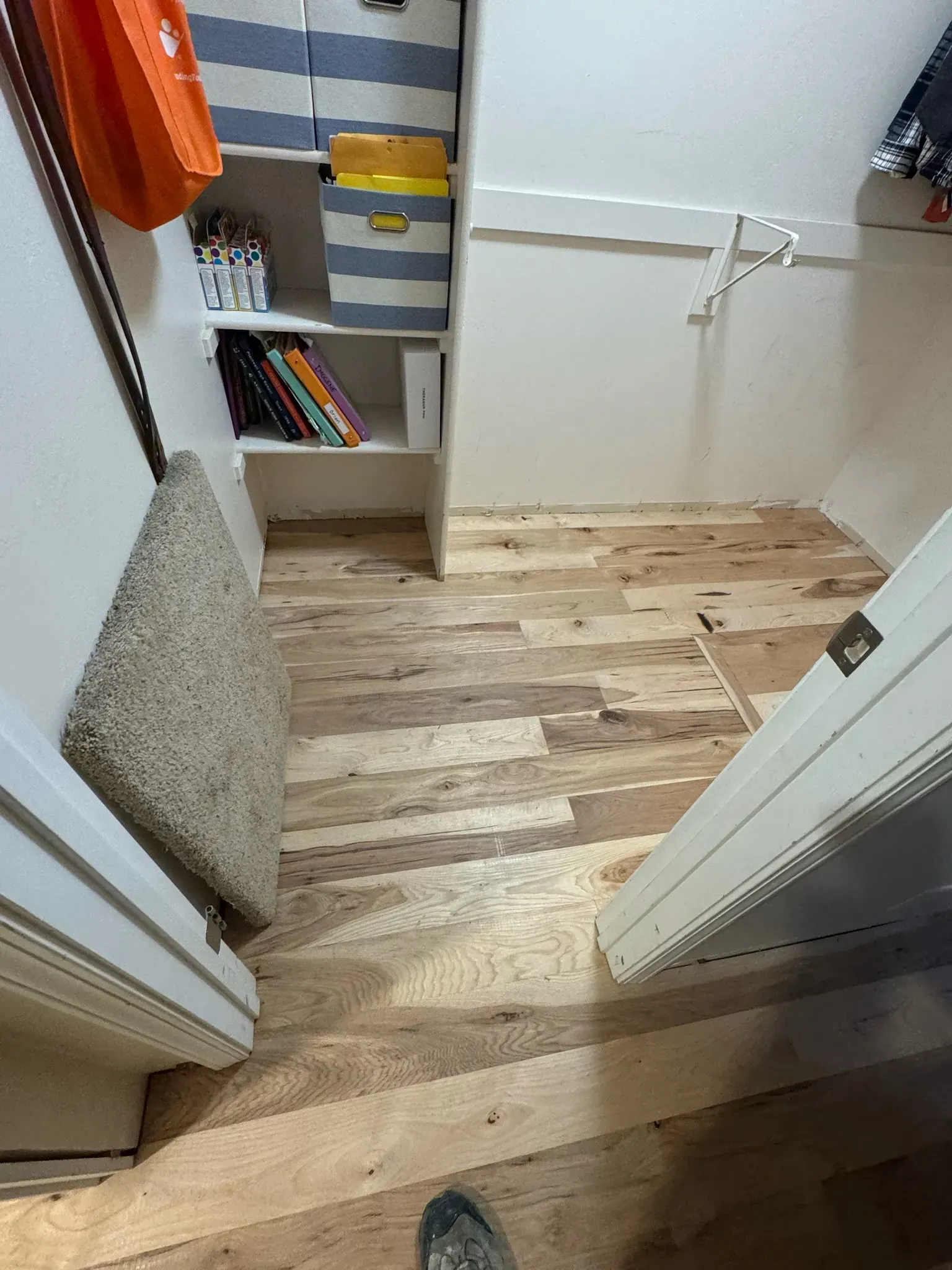 Narrow hallway with wood floor, open doors, and a small white shelving unit with books and folded items.