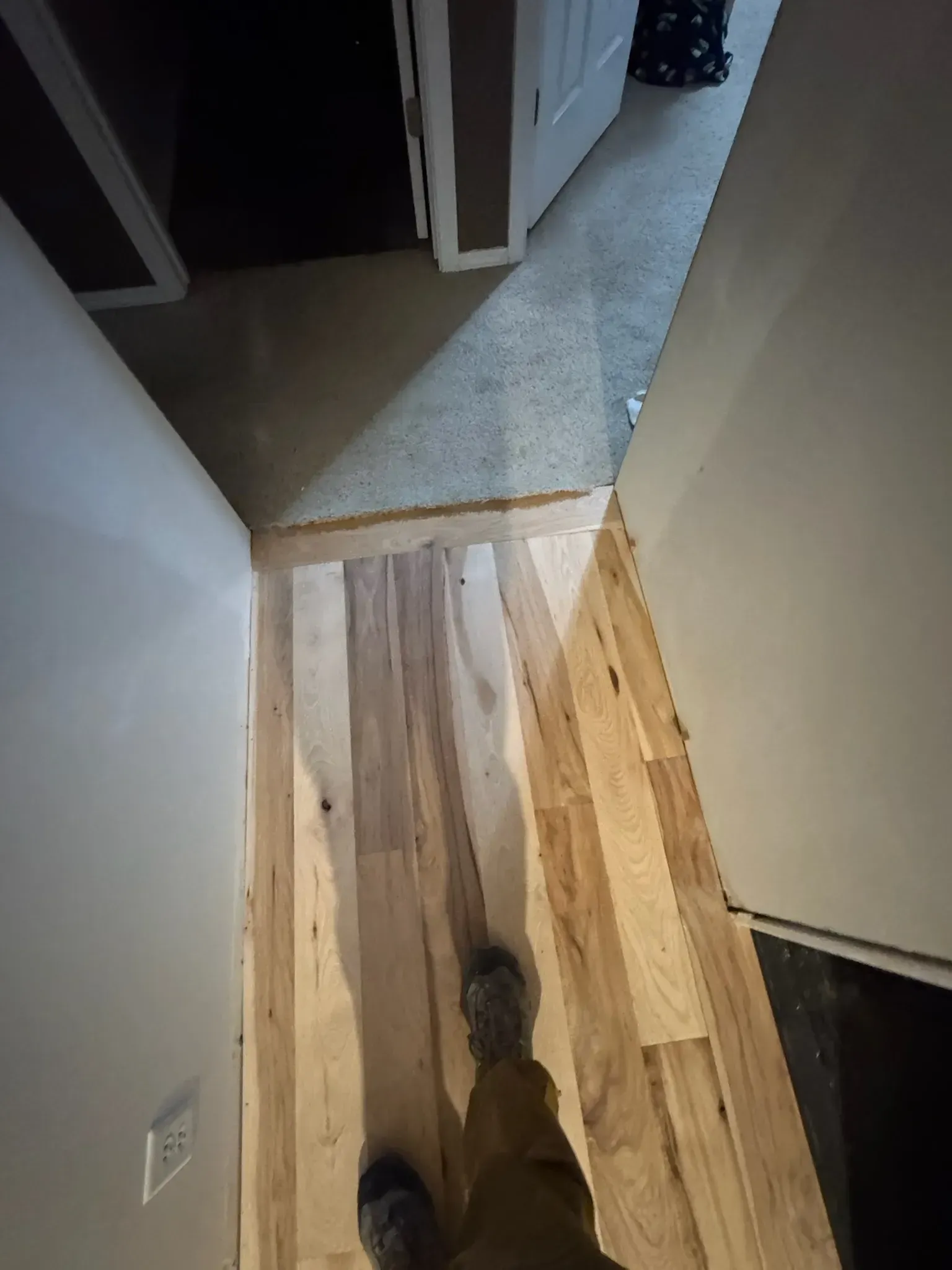 Person’s view of a narrow hallway with light wood flooring and doorways leading into a darker room.