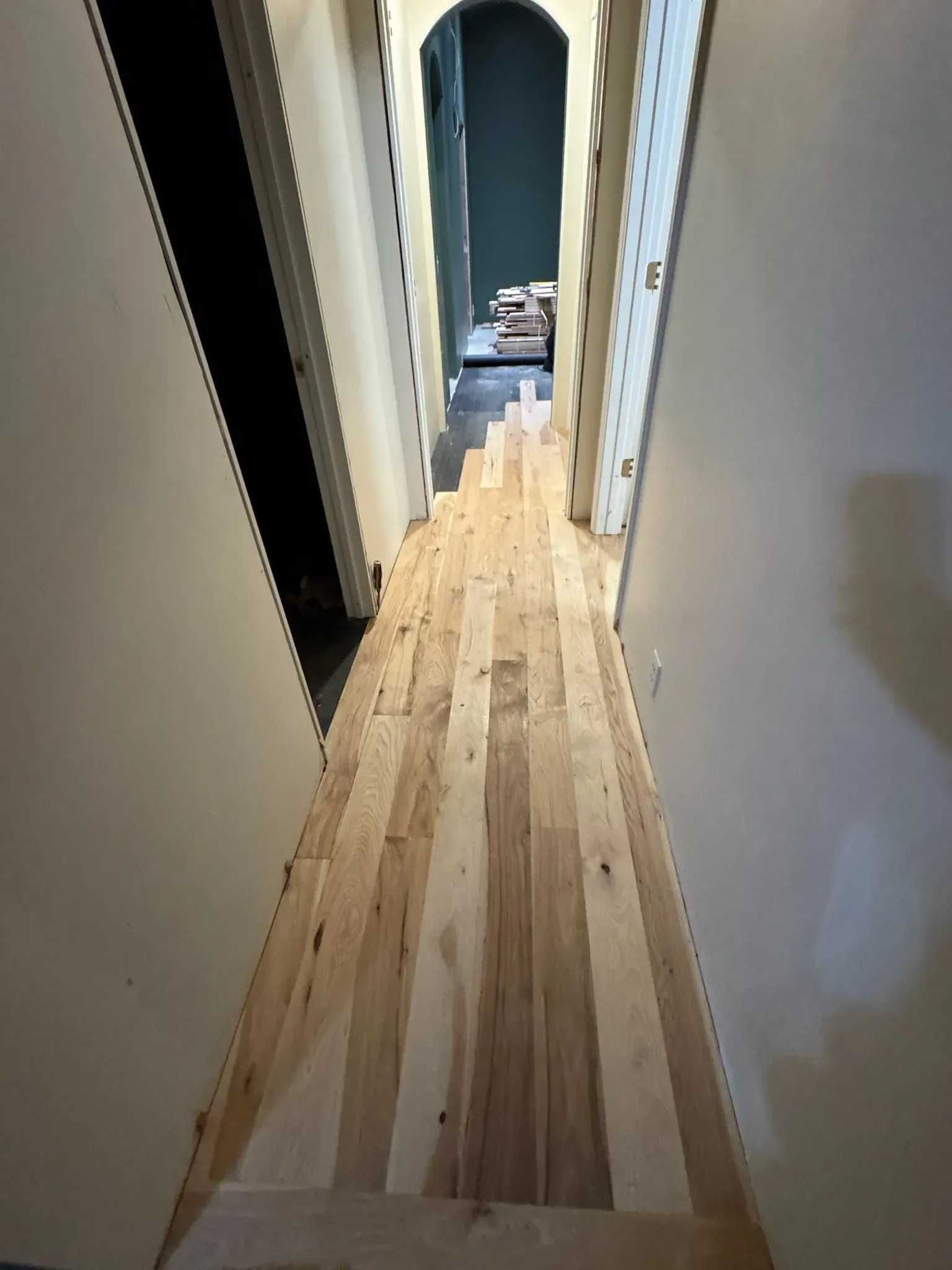 Narrow hallway with light wood flooring and white walls, leading to a dark doorway at the end