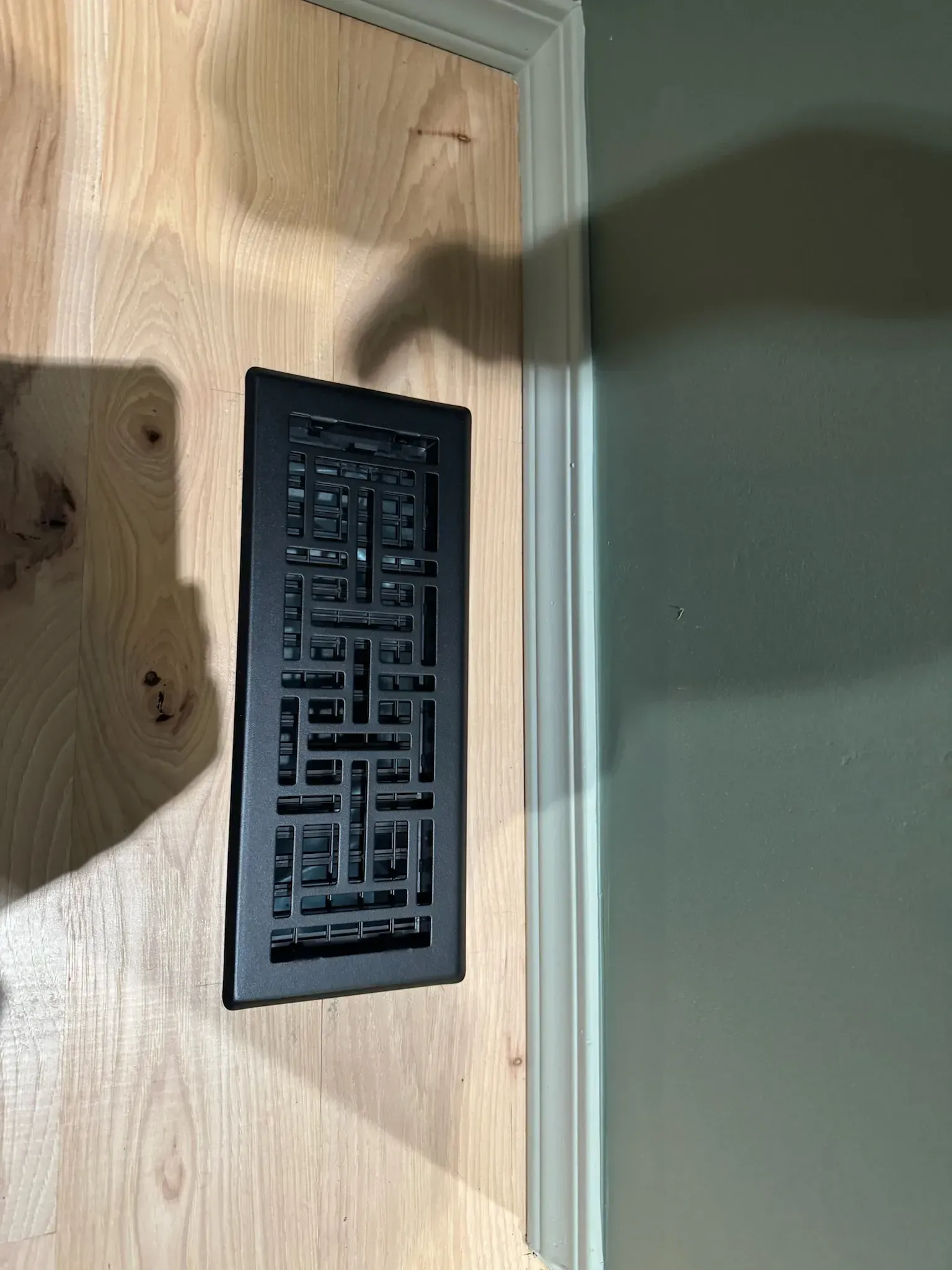 Black rectangular wall vent on a light wood wall with green trim and shadow cast across it