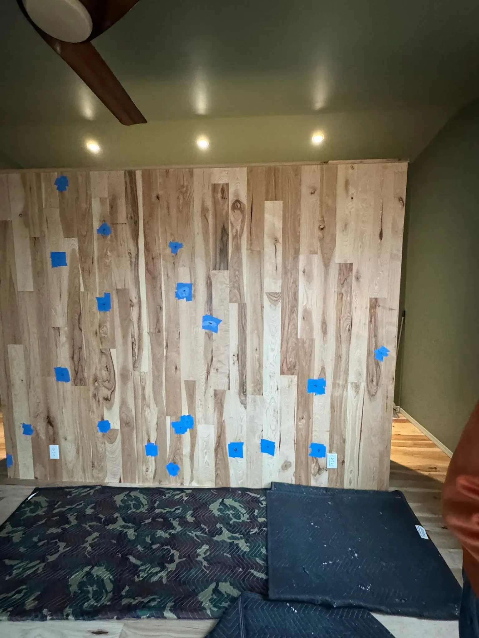 Small room with a wood-paneled wall marked by blue tape and a patterned mattress on the floor.