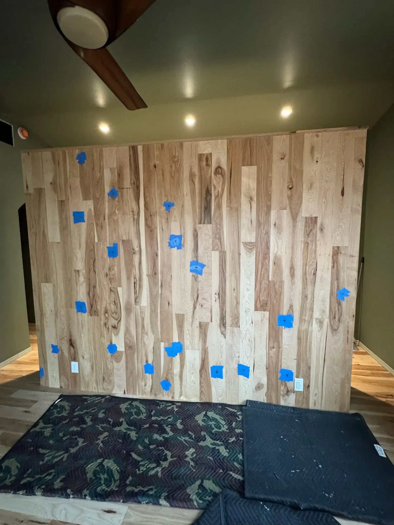 Indoor rock-climbing wall with blue holds above padded mats and a ceiling fan above