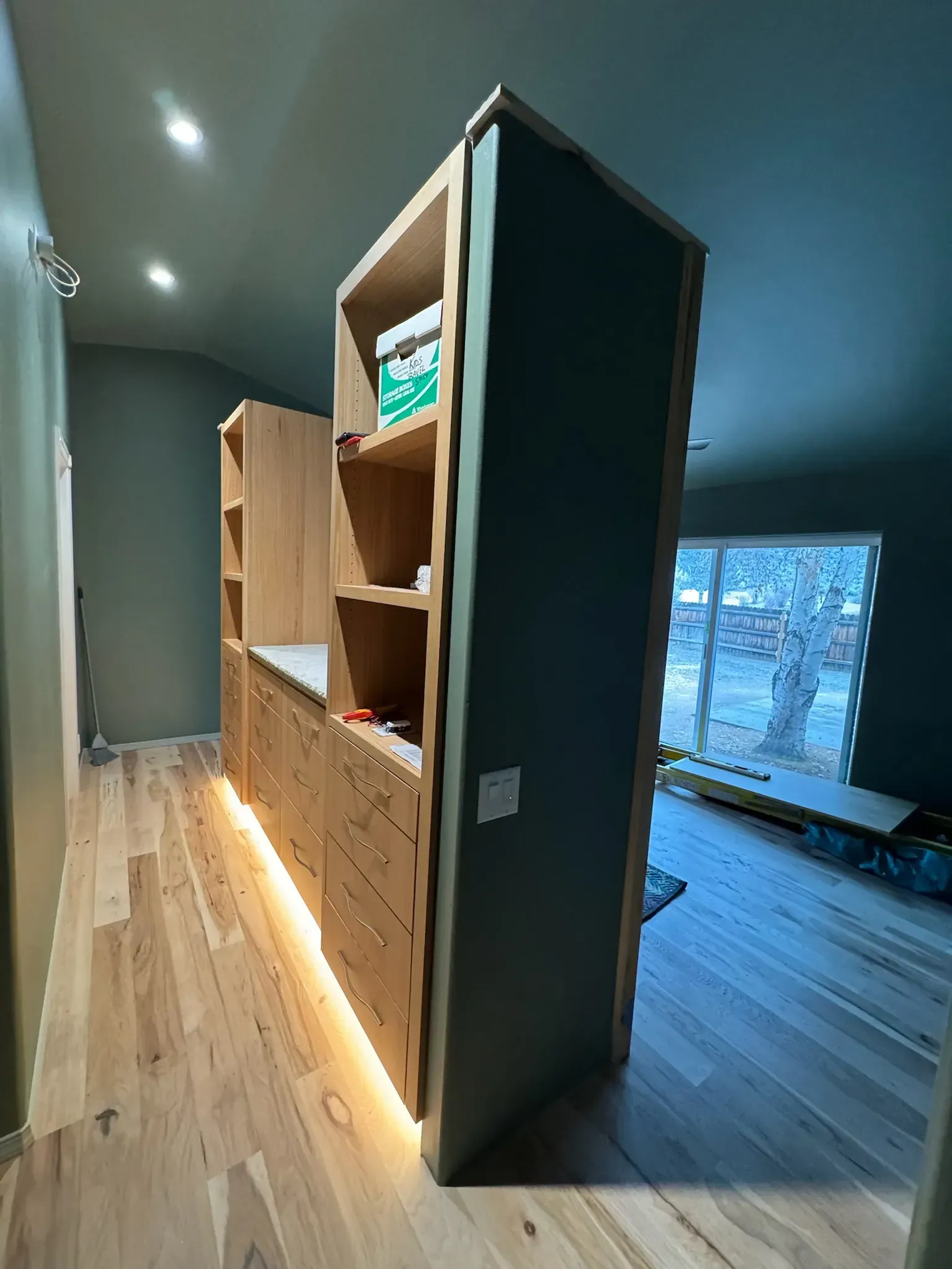 Tall wooden shelving unit with built-in drawers in a modern room, lit by warm floor lights near a window.