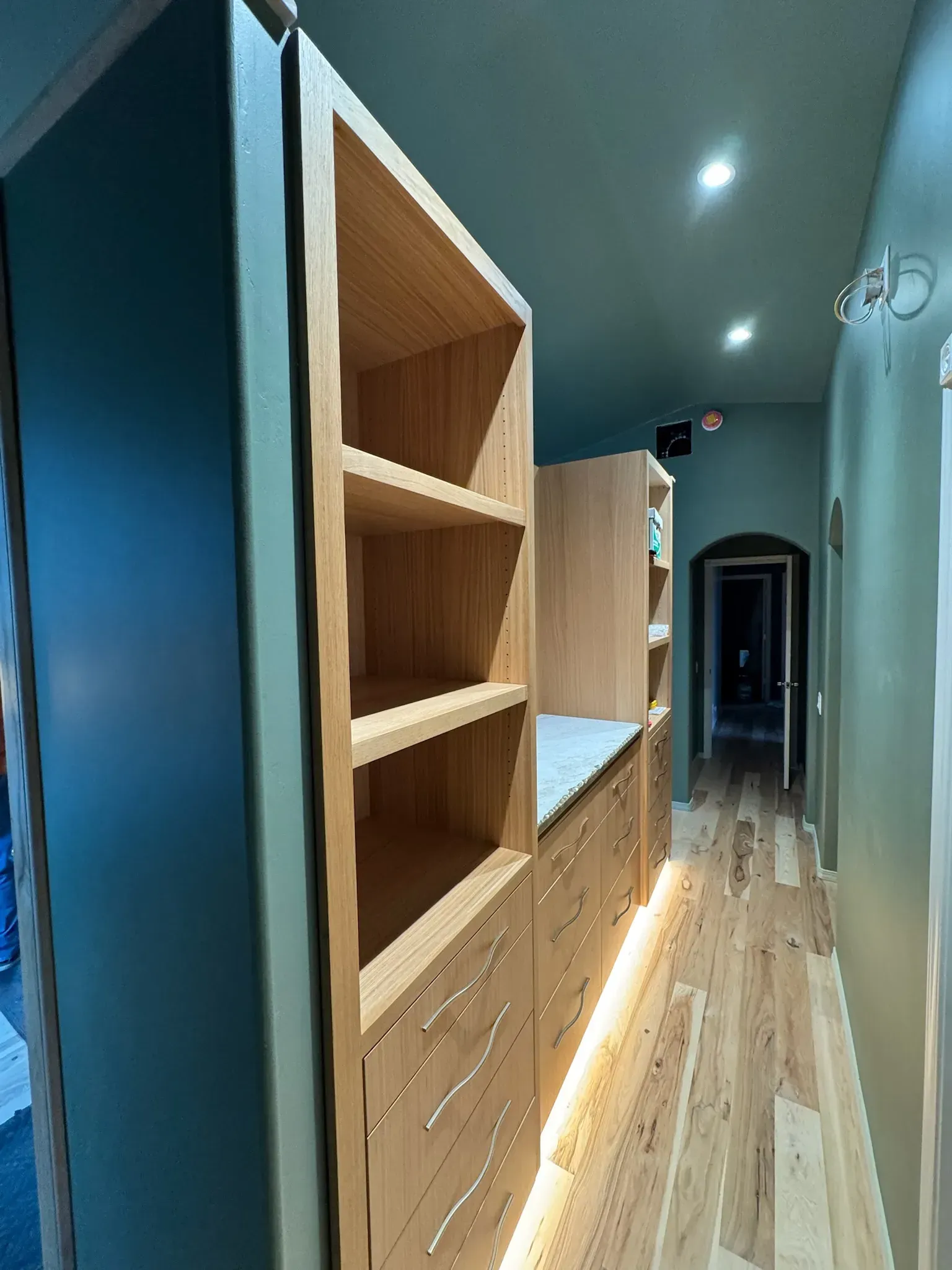 Modern narrow hallway with wooden shelving, cabinets, recessed lighting, and a long wood floor corridor