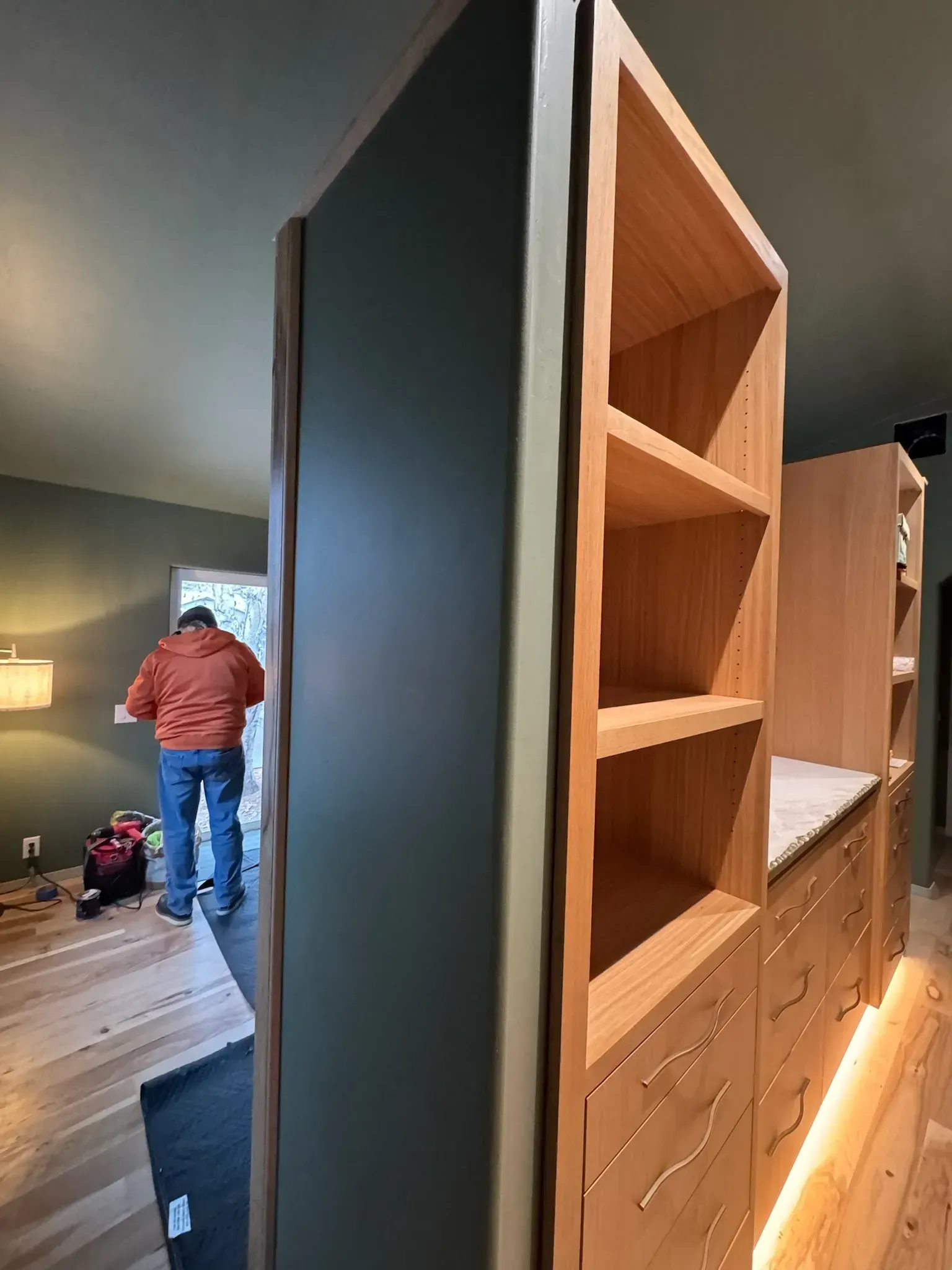 Modern bedroom with built-in wooden shelving and drawers, viewed past a person in a red jacket near the doorway