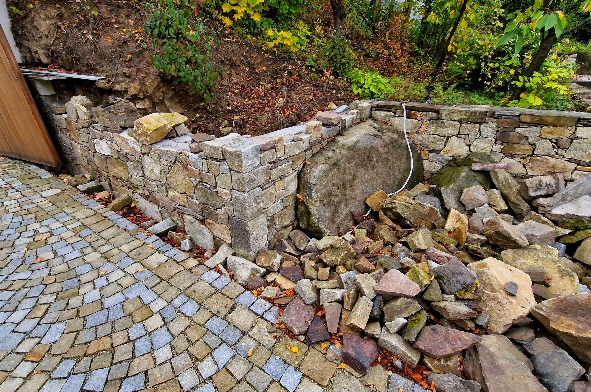 Cobblestone path beside a collapsed stone retaining wall with scattered rocks and autumn foliage