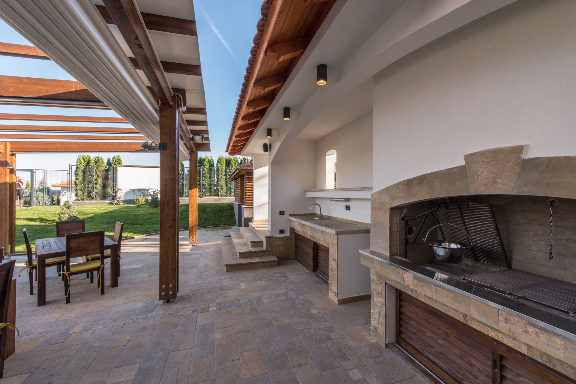 Covered outdoor patio with stone fireplace, built-in grill, and dining table under wooden beams