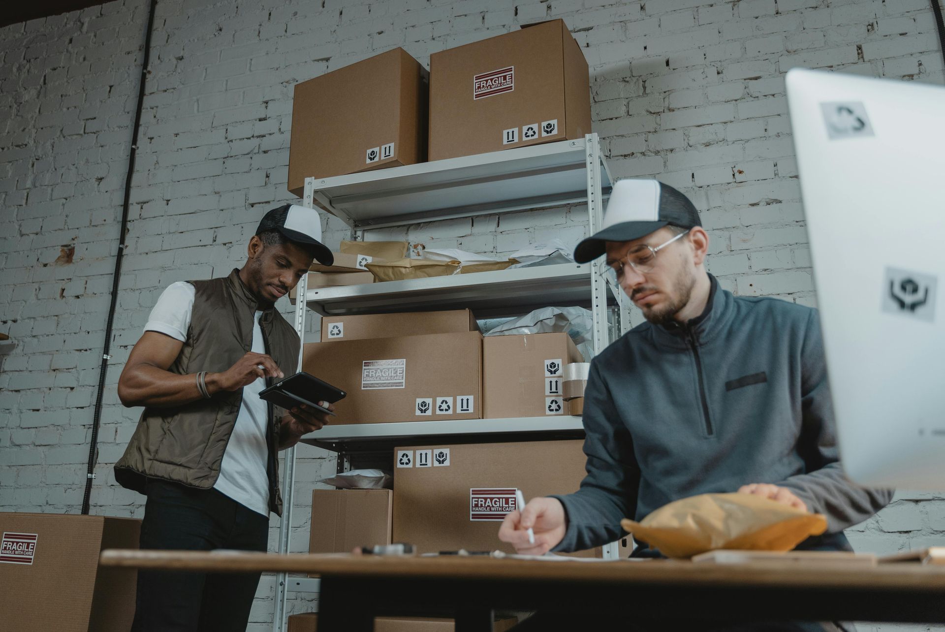 Two people working in a warehouse, one on a tablet, the other at a computer, surrounded by boxes.