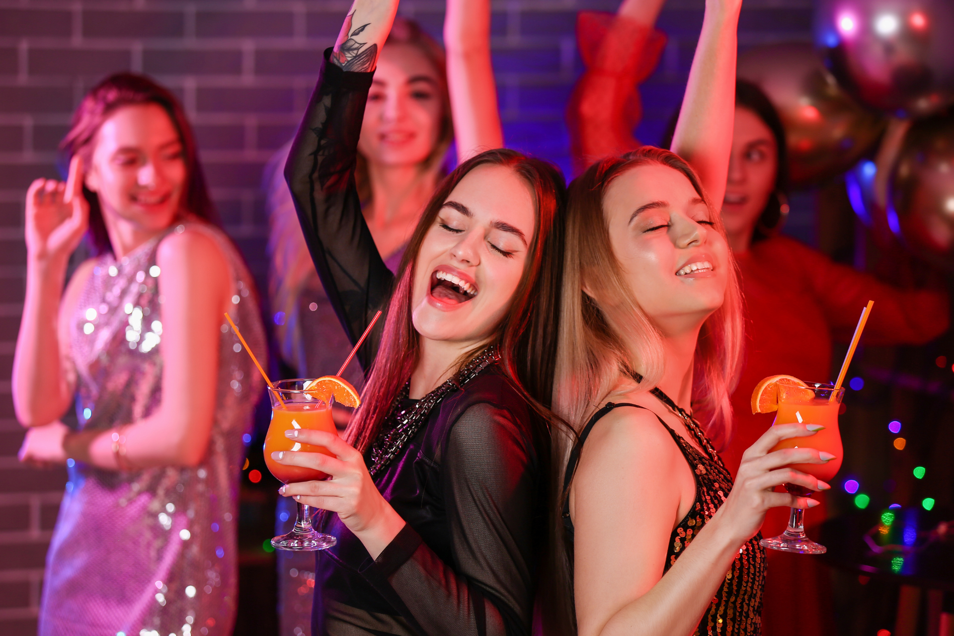 Friends dancing and smiling at a colorful party, holding drinks and celebrating together