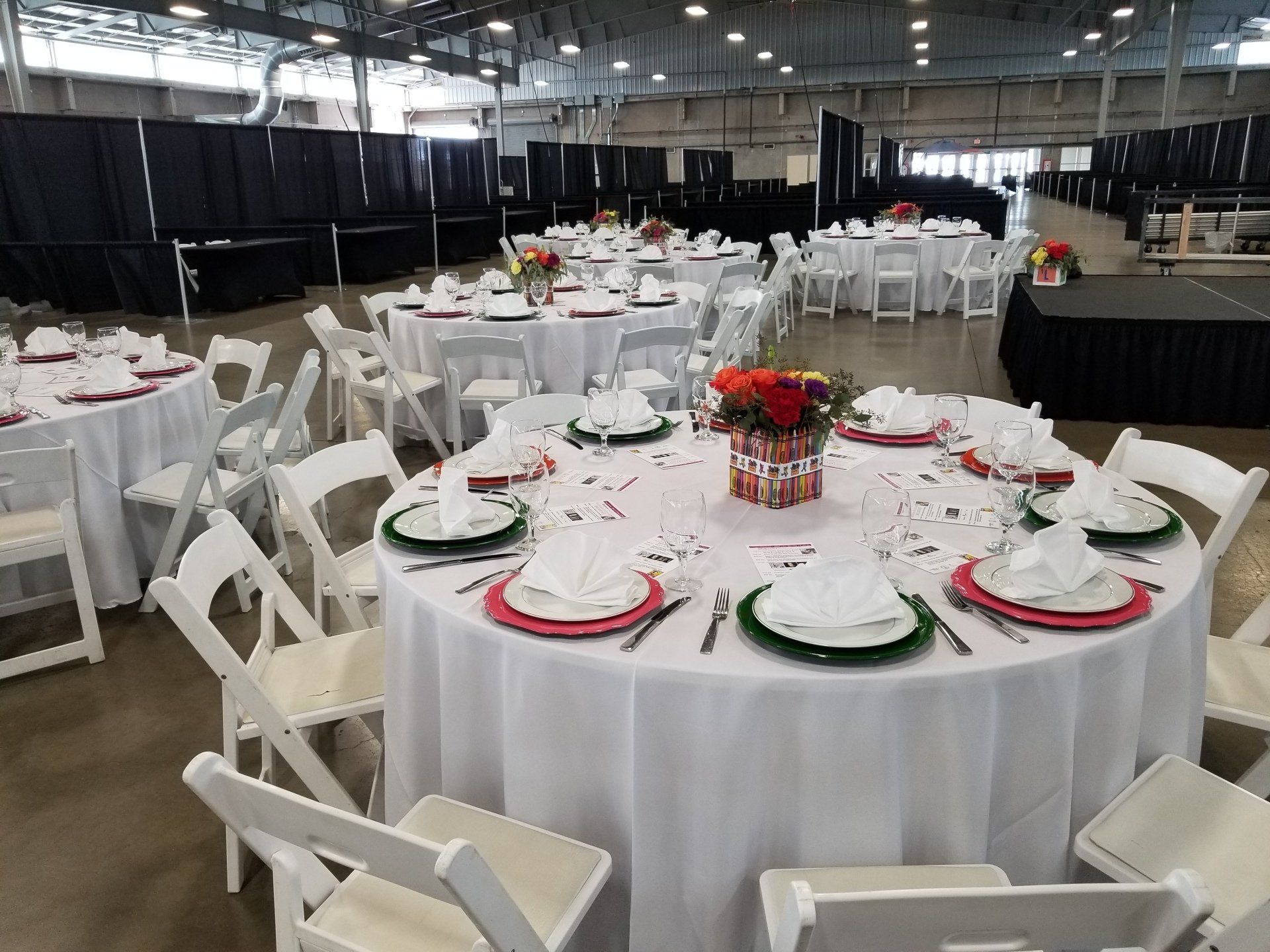 Round tables set for an event with white tablecloths, chairs, and colorful place settings, inside a large venue.
