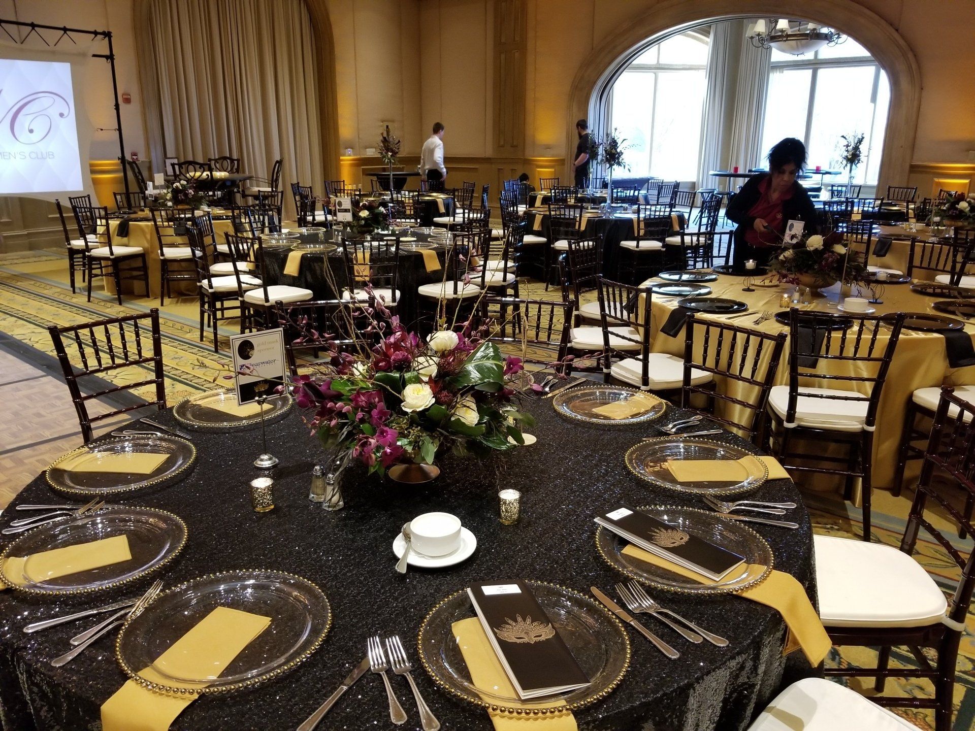 Formal dining room set for an event with black tablecloths, gold accents, and floral centerpieces.