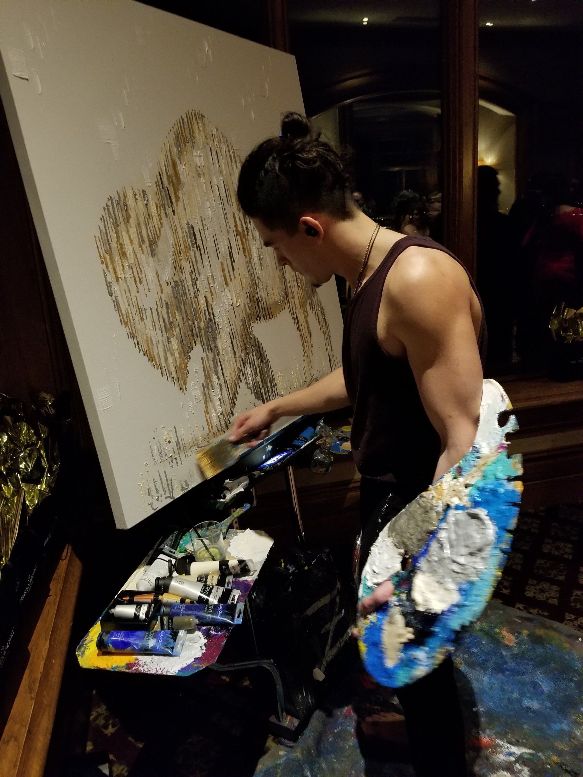 Artist painting with brush, standing before a large canvas. Palette of paints in hand; work surface nearby.