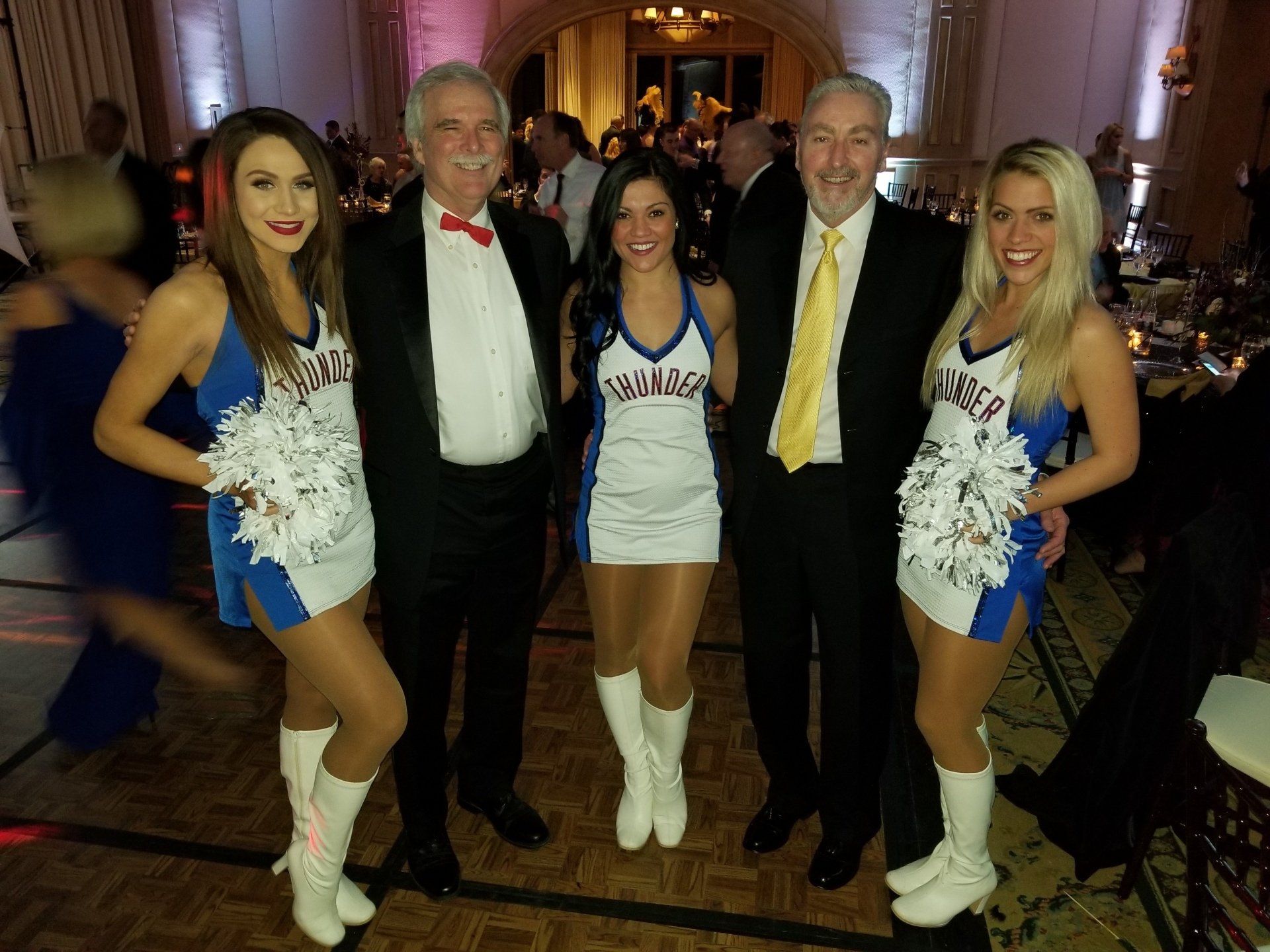 Two men pose with three cheerleaders in formal attire at an event, indoor setting.