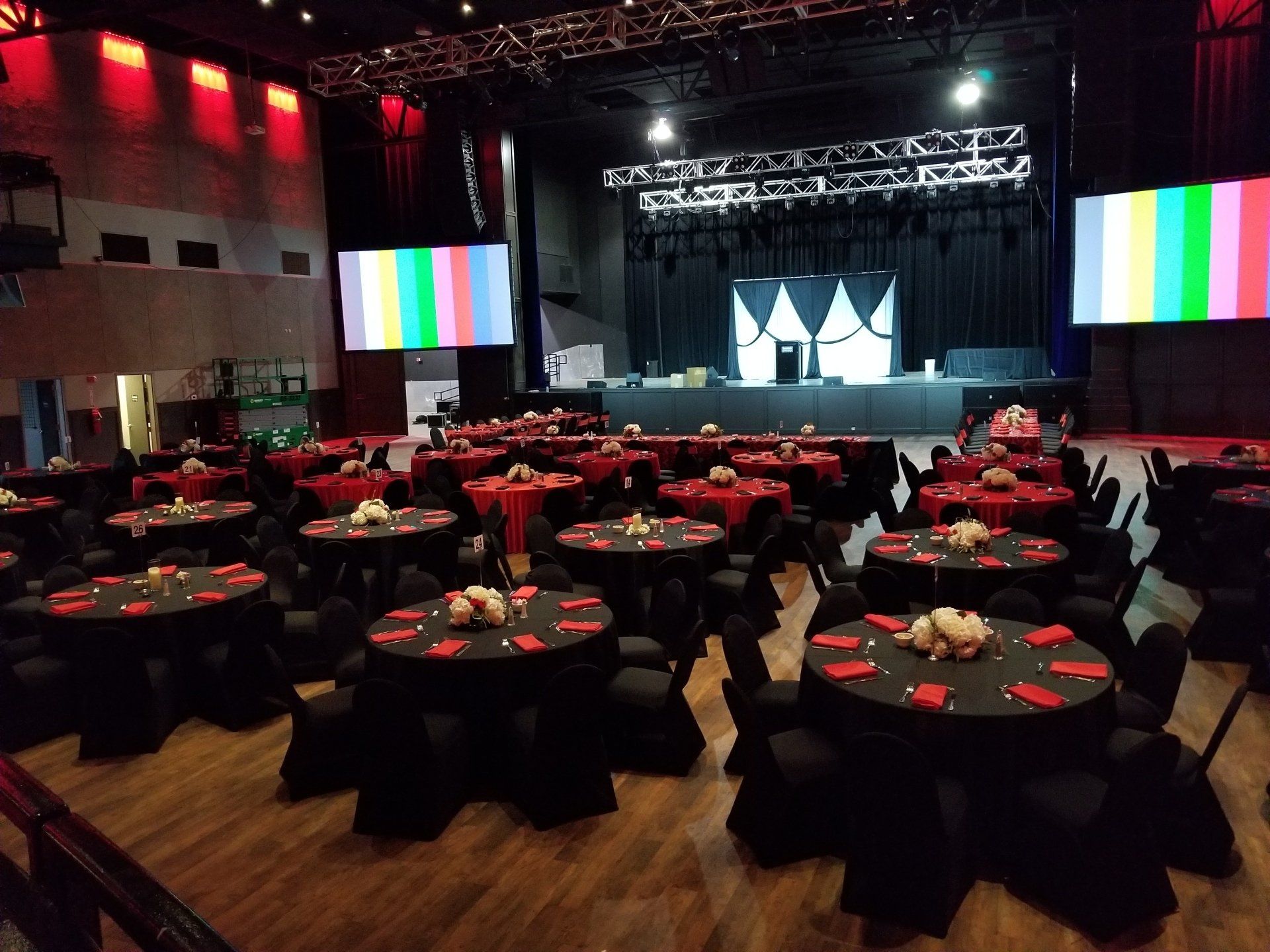 Tables set for an event on a stage with screens displaying color bars. Black table cloths, centerpieces, and stage lighting.