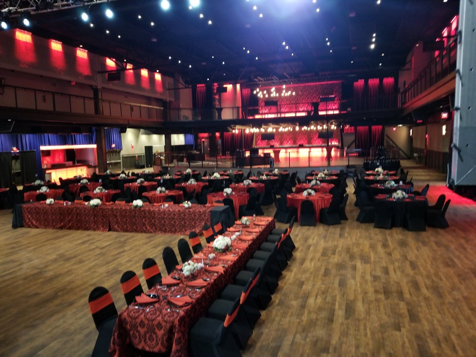 Large event space set up for a dinner. Red and black tablecloths and chairs. Stage in the background.