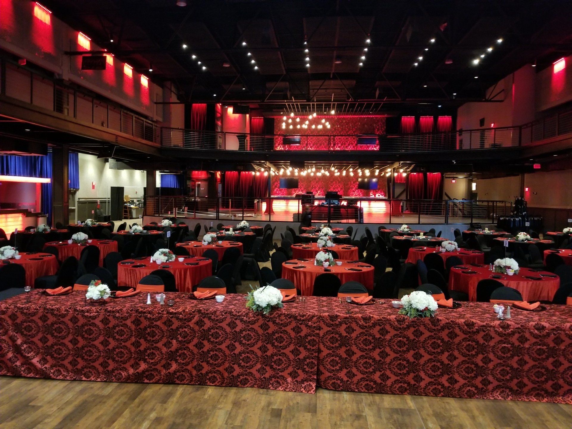 Banquet hall set for event, red and black decor, tables with centerpieces, stage in background.