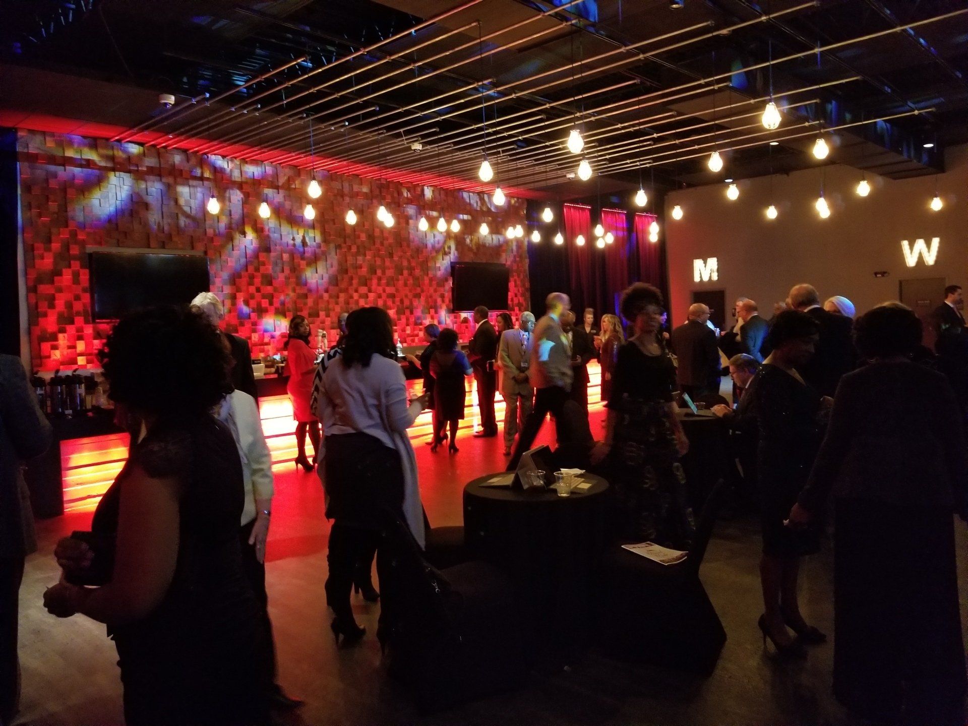 People gather at a lit event space. Red and warm white lighting illuminates walls and the crowd.