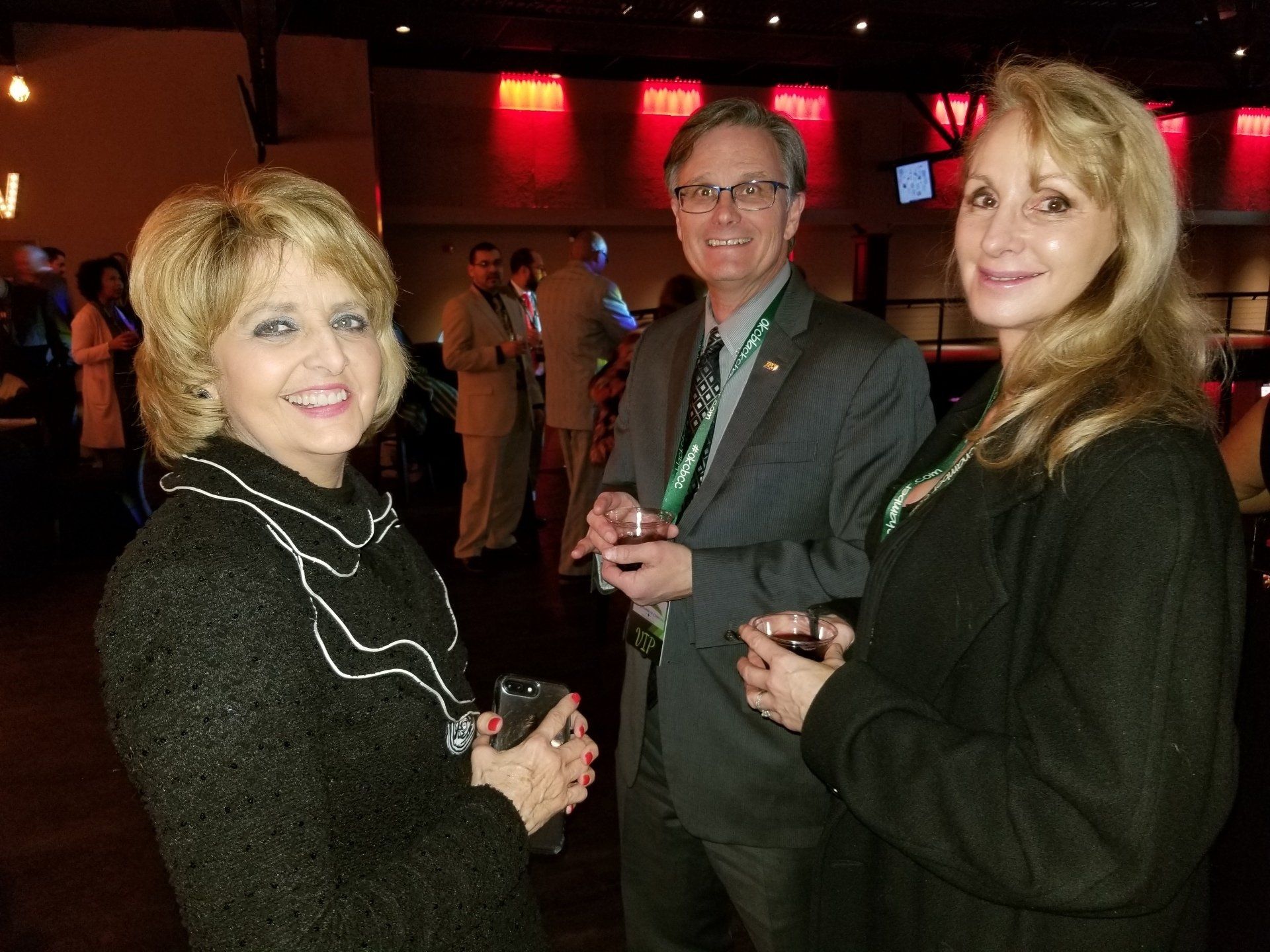 Three people smiling, holding drinks, in a dimly lit room with red accents, likely a social event.