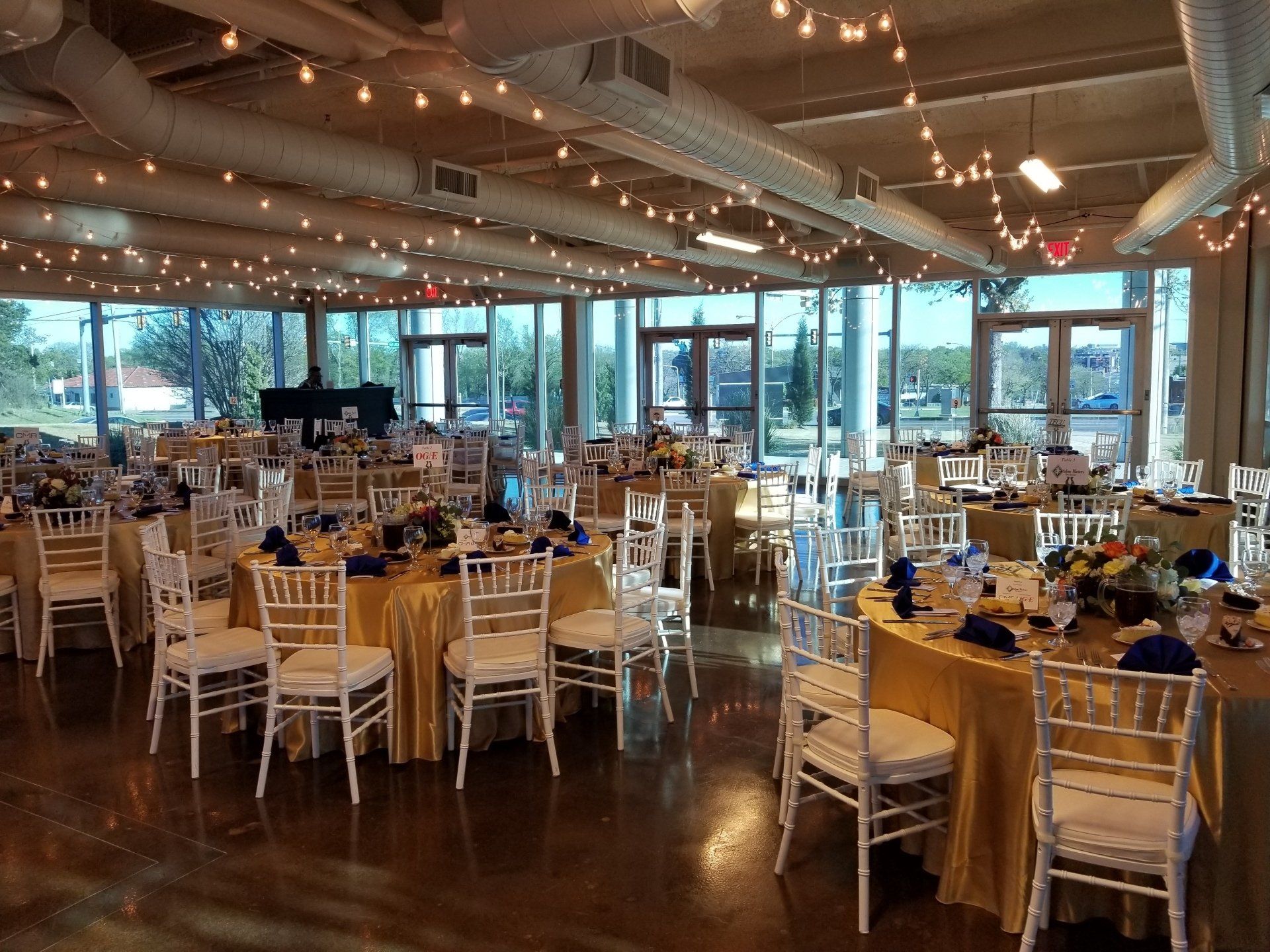Event space with round tables set for guests, gold tablecloths, white chairs, and string lights.