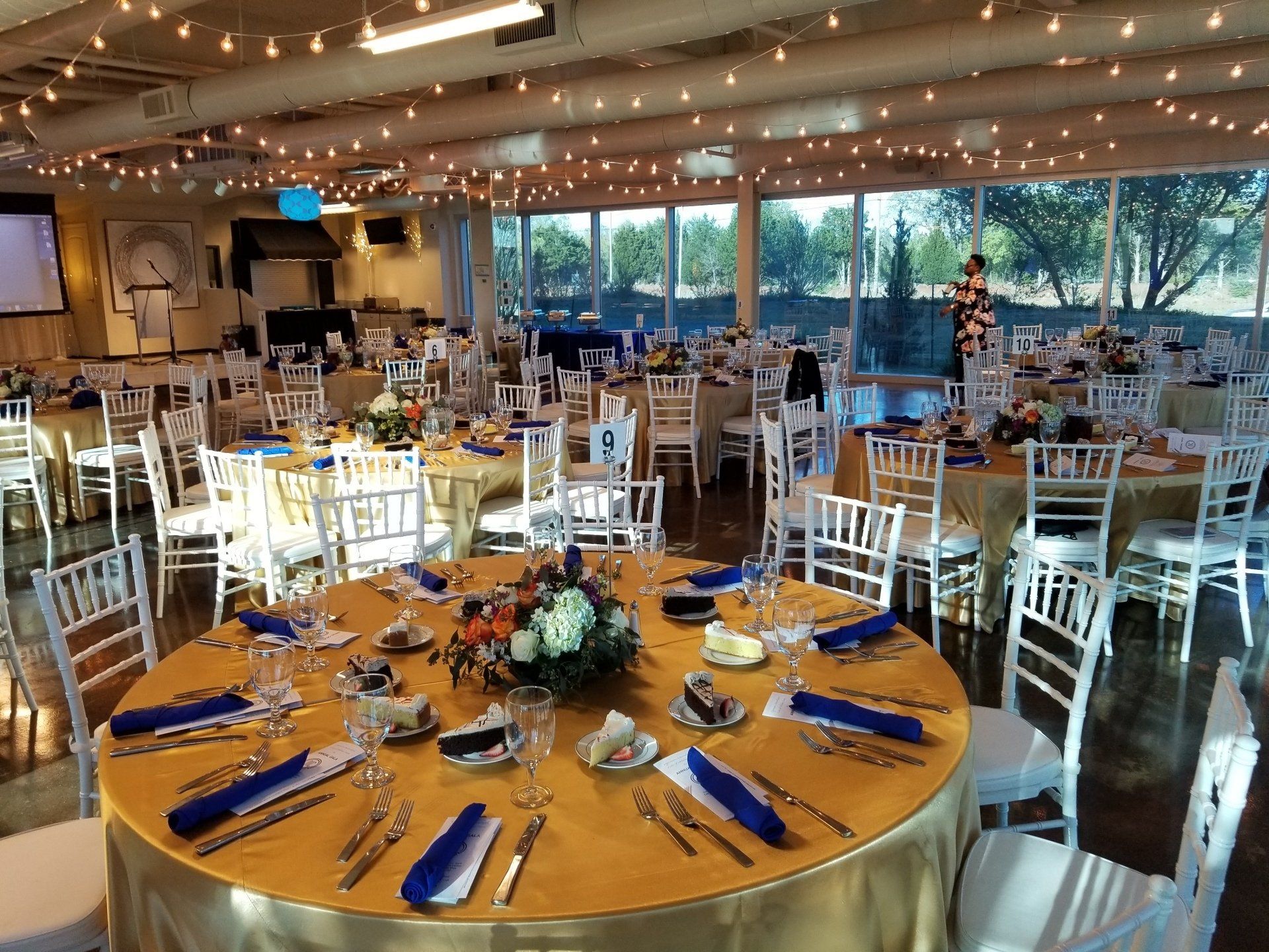 Banquet hall set with gold tablecloths, blue napkins, and floral centerpieces. Bright with string lights and outdoor view.