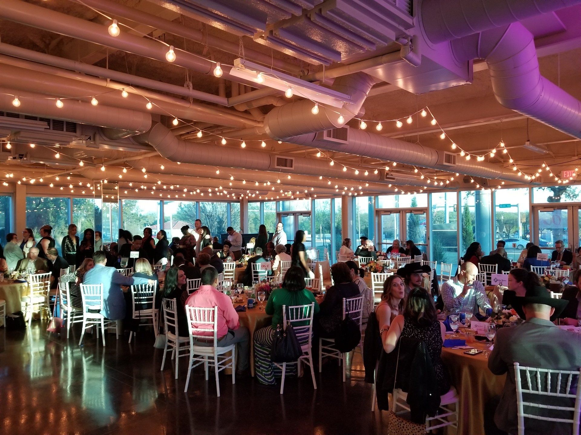 Large event space with people seated at tables, warm lighting, and string lights.