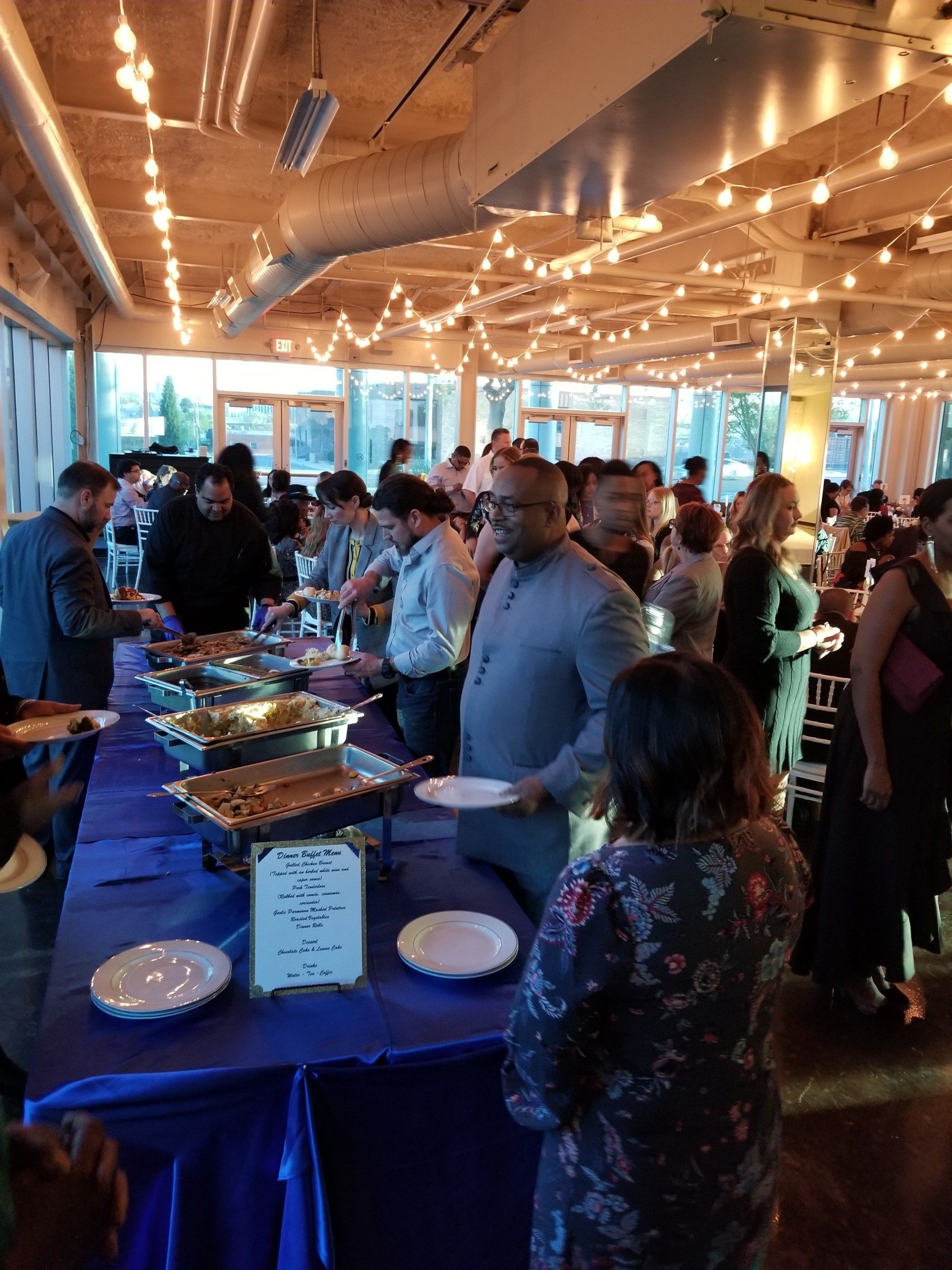 Buffet at event with people serving food from tables. Blue tablecloth and string lights.