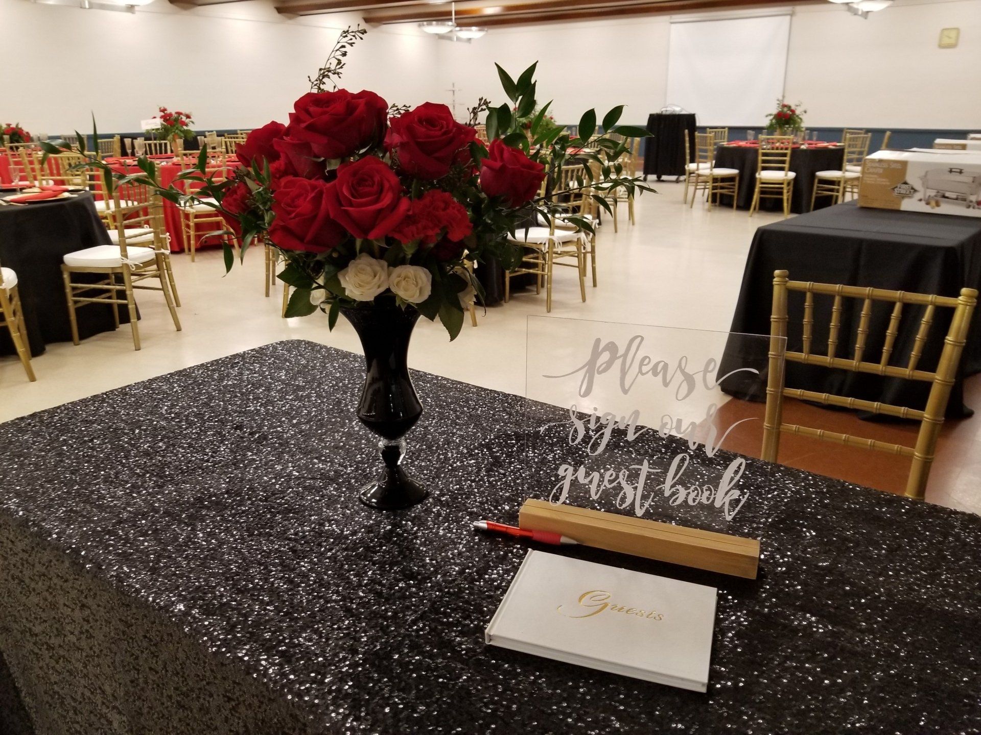 Reception hall with tables, gold chairs, red roses, guest book, and sign.