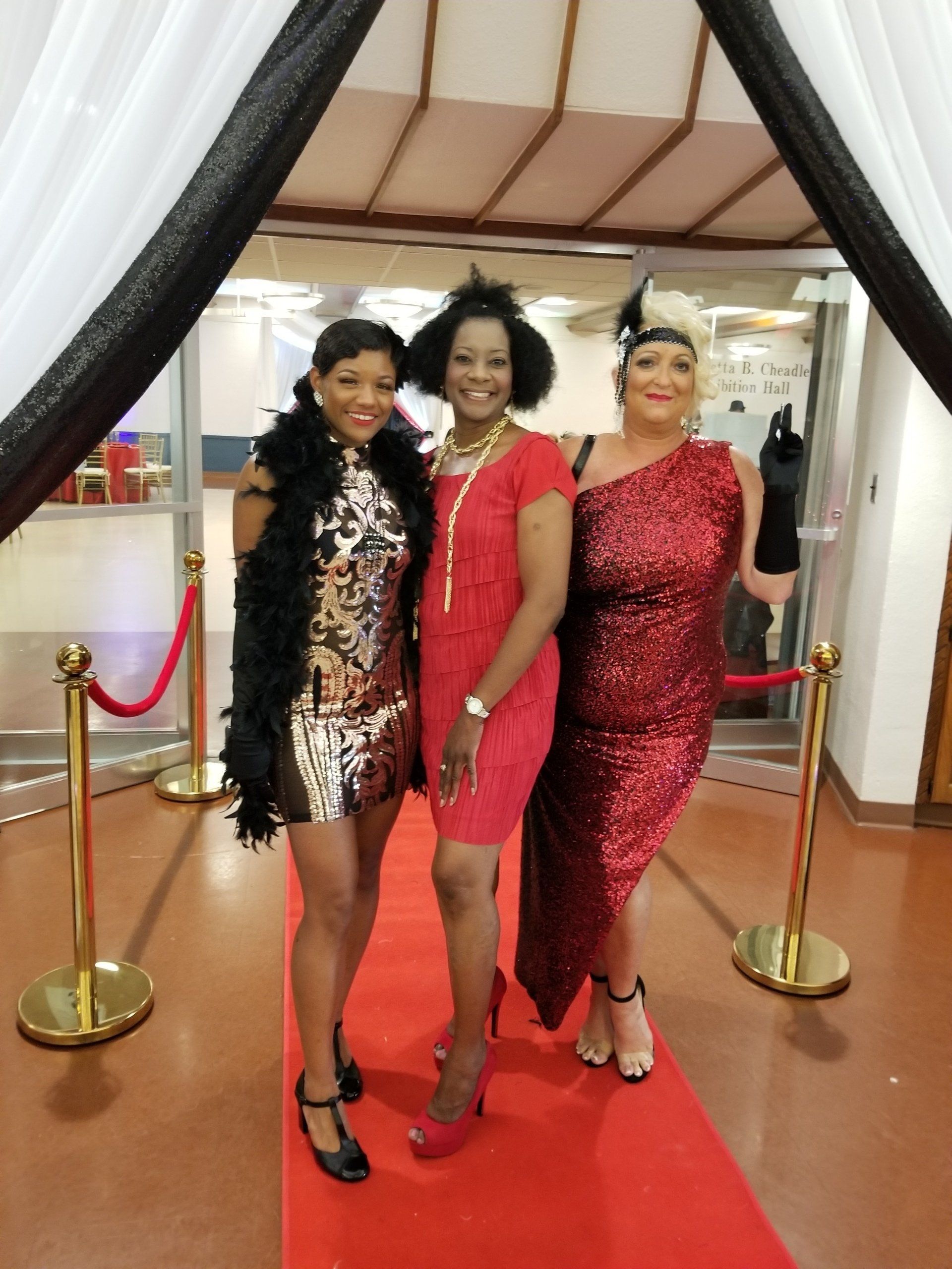 Three people in flapper dresses pose on a red carpet with black and gold decor.