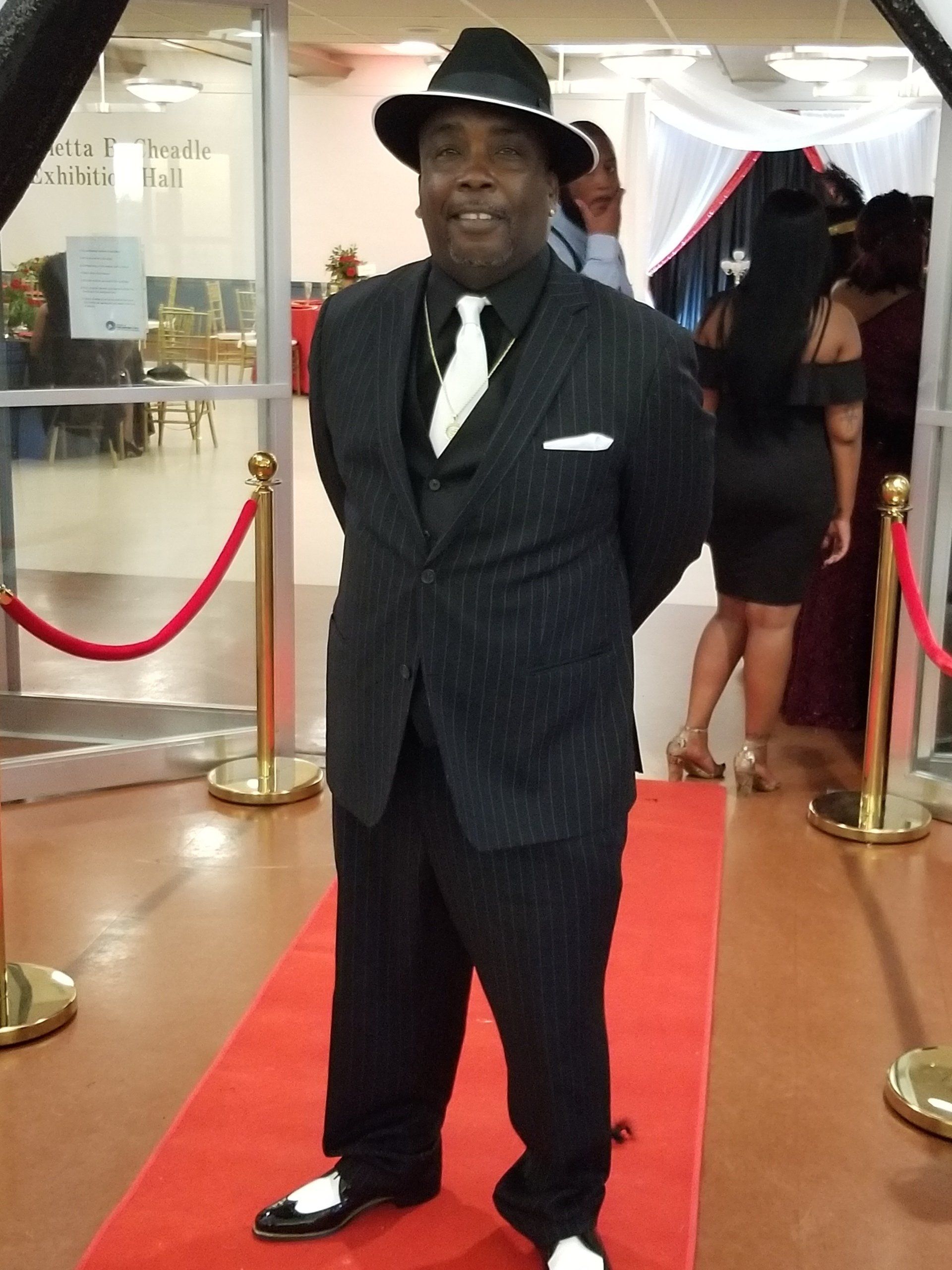 Man in black pinstripe suit and fedora poses on a red carpet.