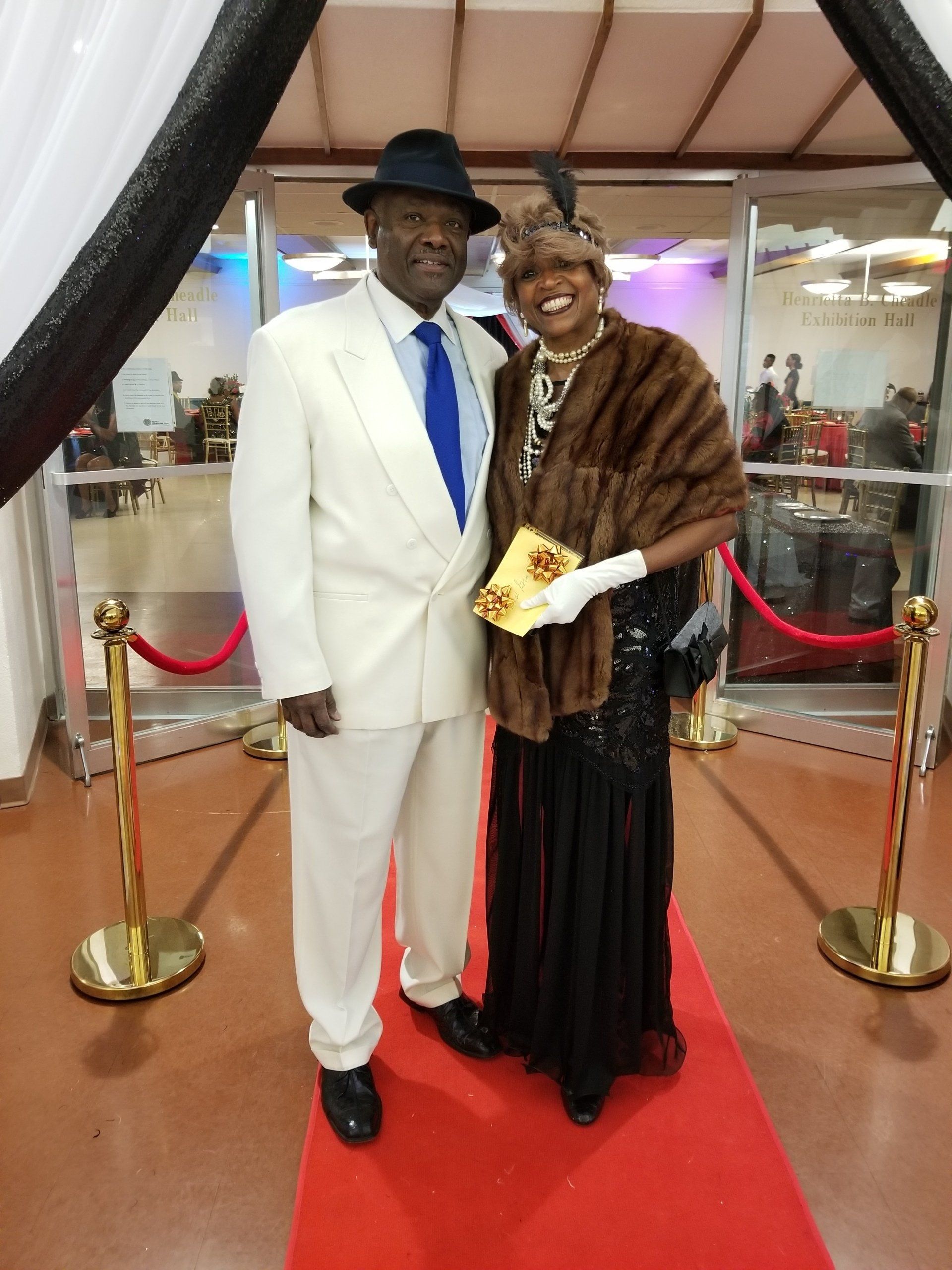 Couple in 1920s attire poses on a red carpet. The woman wears a fur stole and the man wears a white suit.