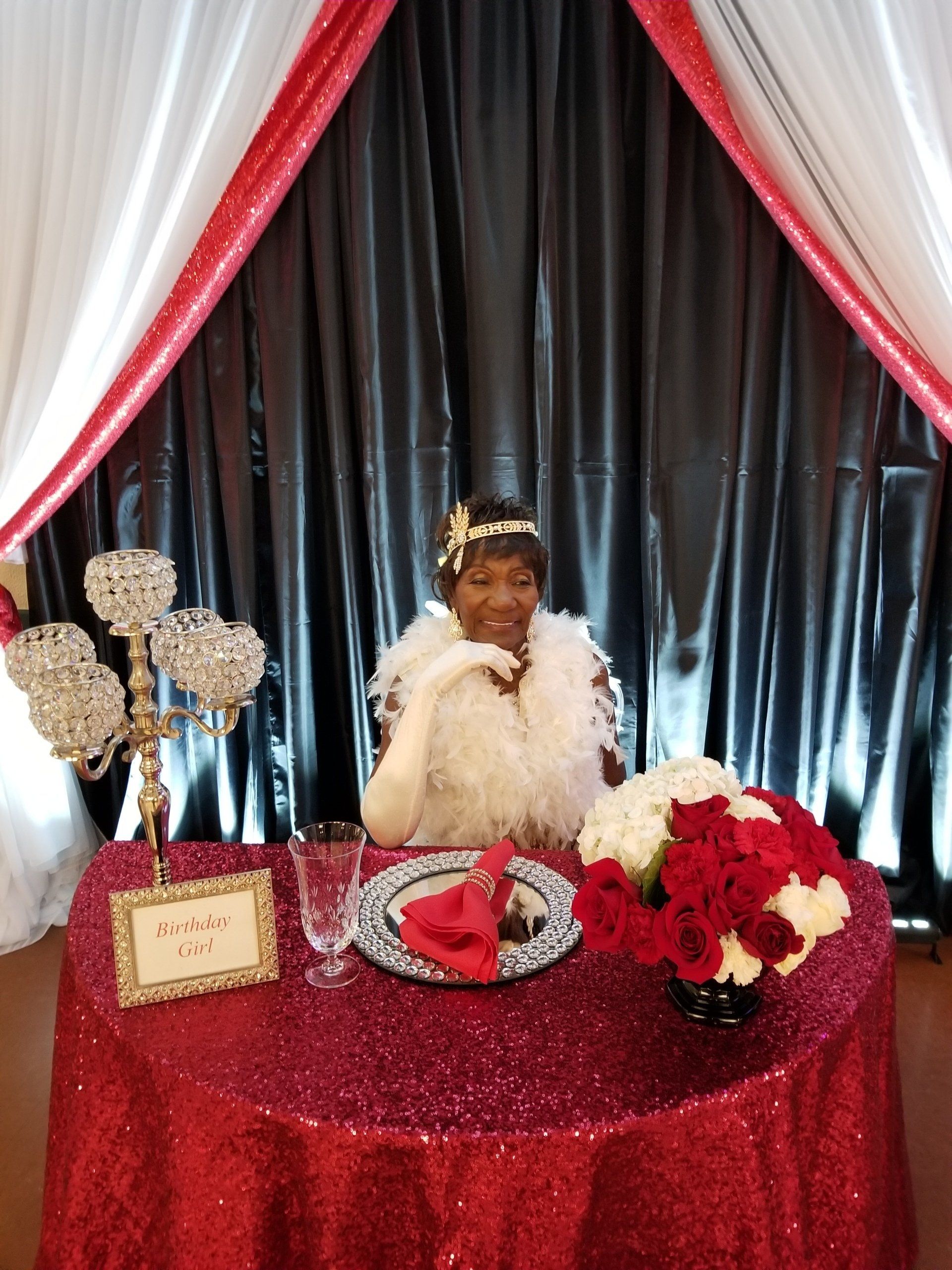 Woman in white fur at red-sequined table. Flowers, crystal candelabra, and backdrop of black, red, and white.