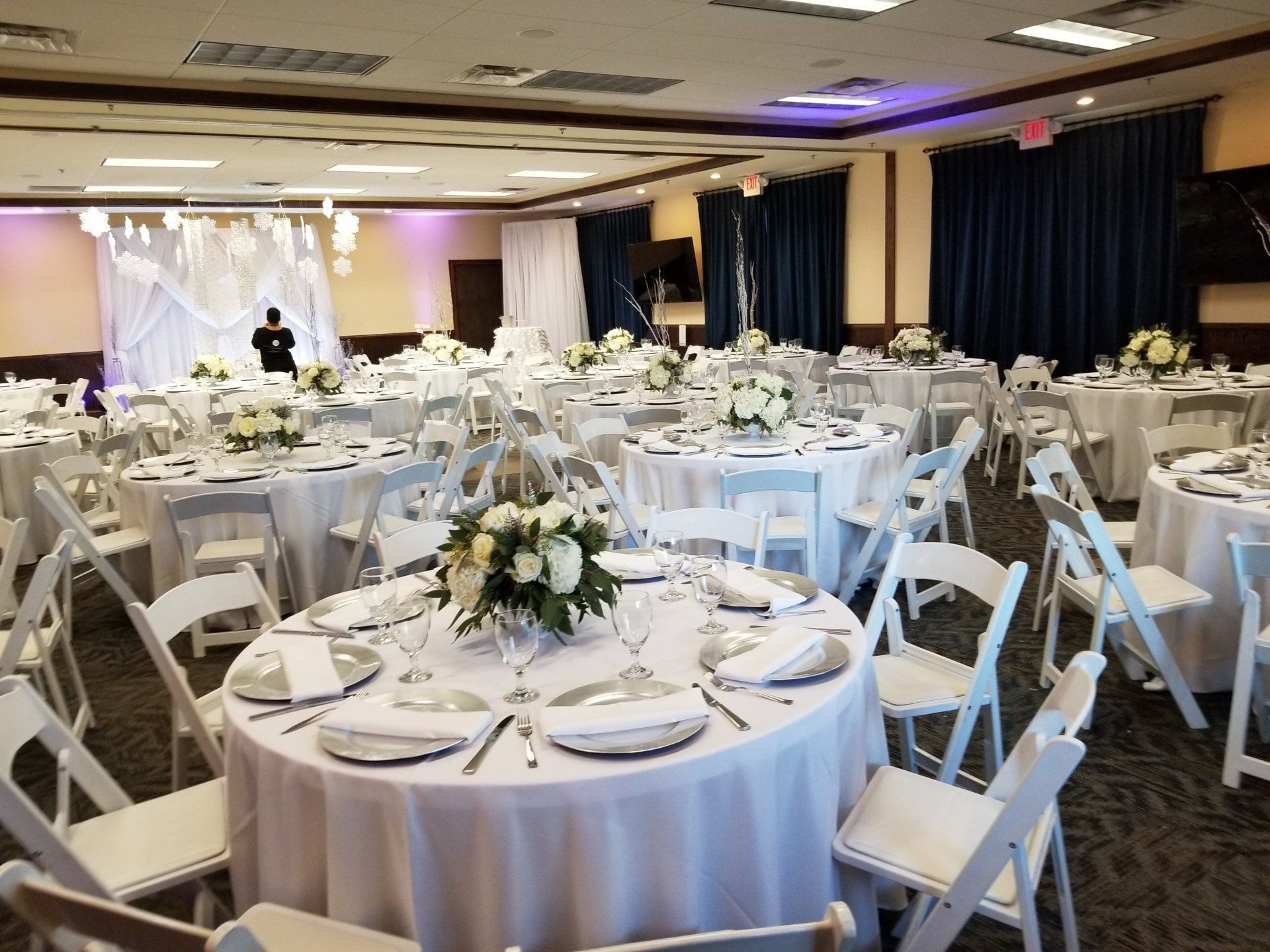 Wedding reception hall with round tables set for guests; white linens, chairs, and centerpieces.