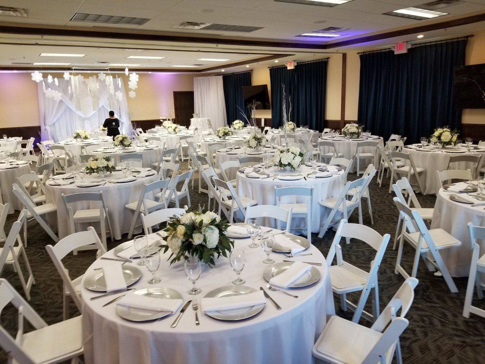 Banquet hall set for an event; round tables with white linens, chairs, floral centerpieces, and place settings.