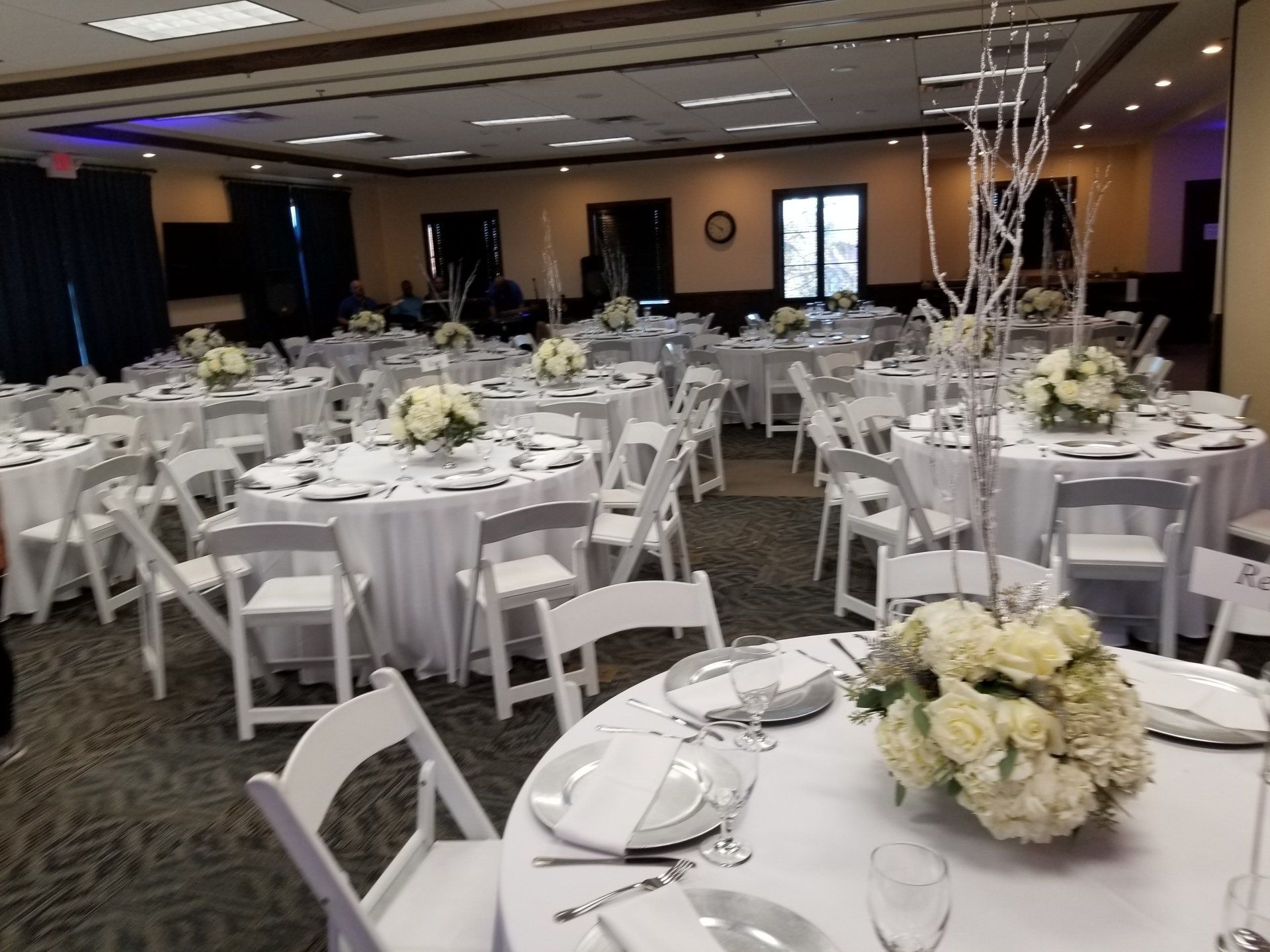 Formal event space with white round tables, chairs, and floral centerpieces; neutral tones.
