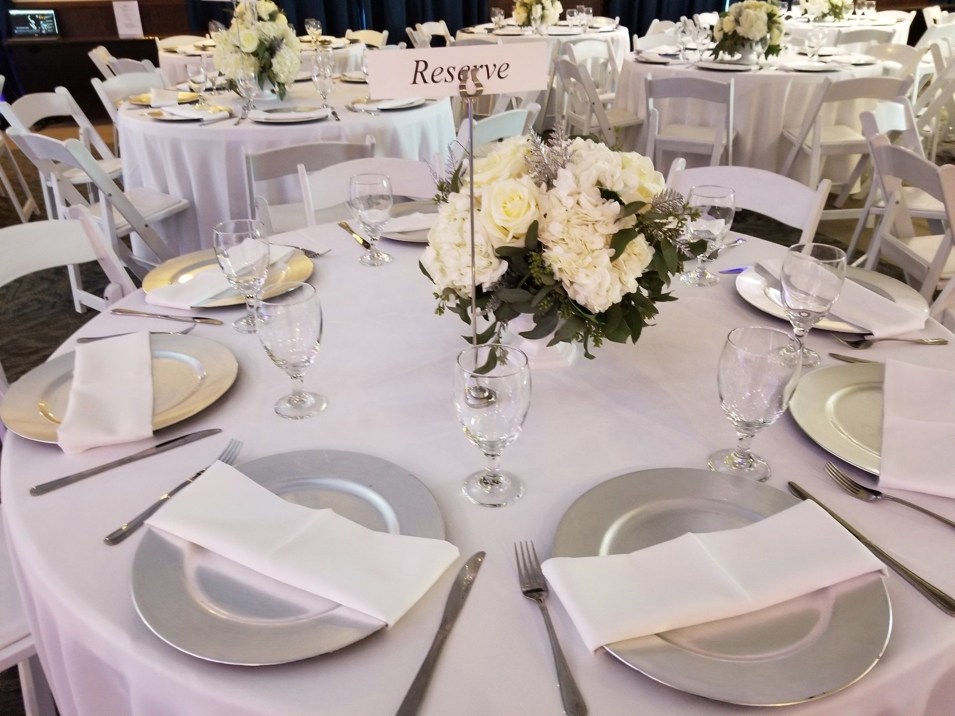 Round table set for a formal event with white linens, silver chargers, flowers, and