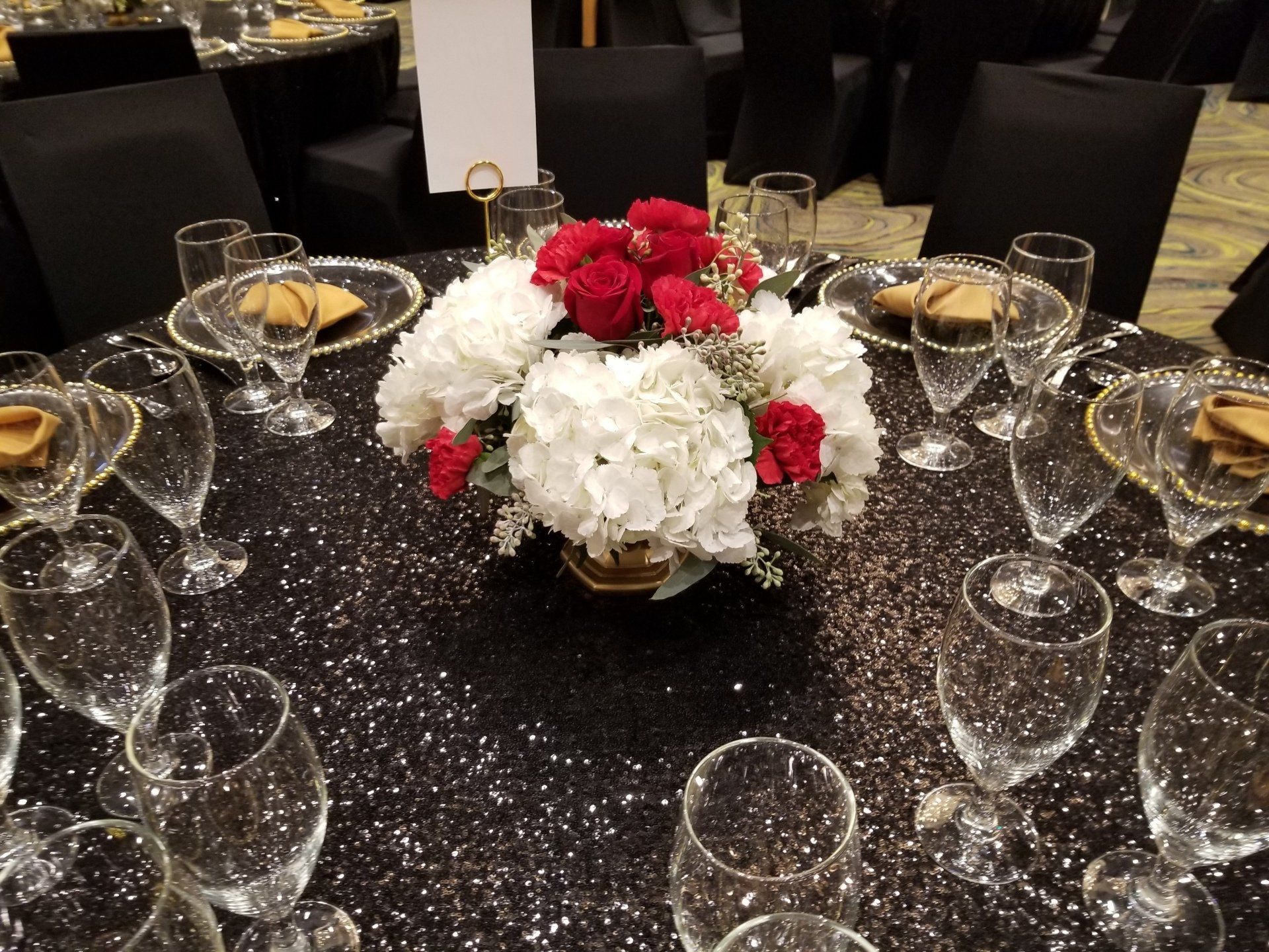 Round table set for a formal event, black sequin tablecloth, floral centerpiece, and glassware.