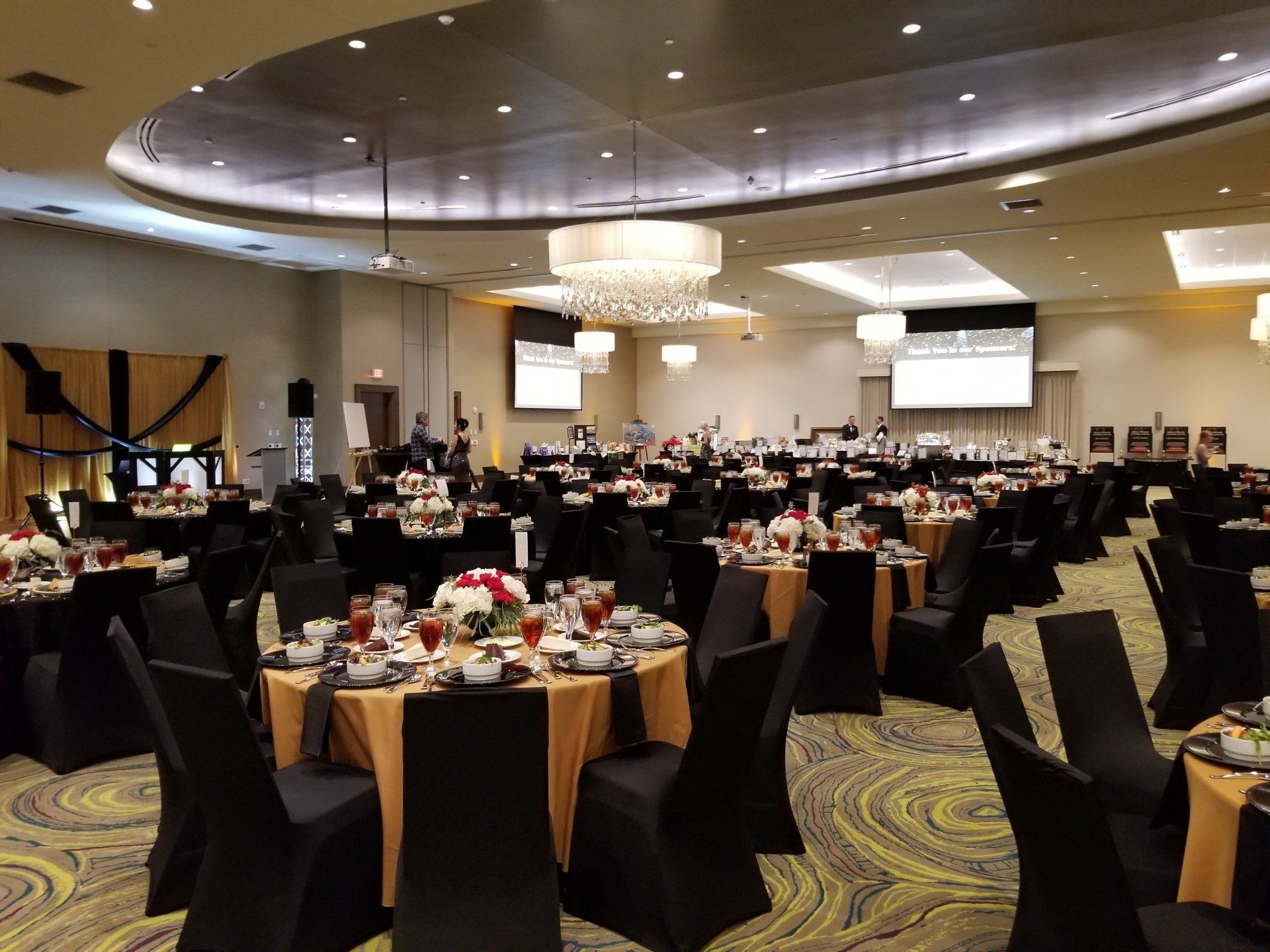 Ballroom set for an event; round tables with gold linens, black chair covers, and floral centerpieces.