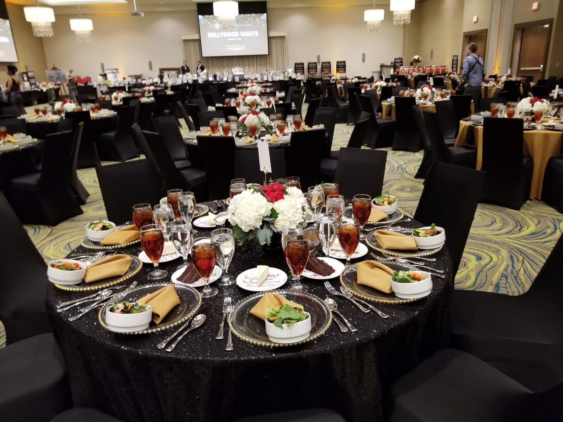 Round tables set for a formal event, black tablecloths, centerpieces, food on plates, people in background.