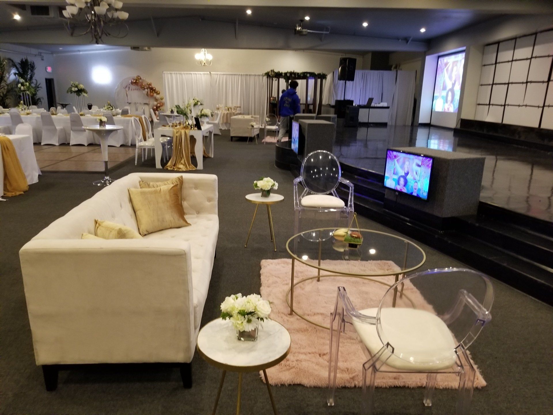 Event space with white sofa, gold accents, tables, and a stage with a TV screen.