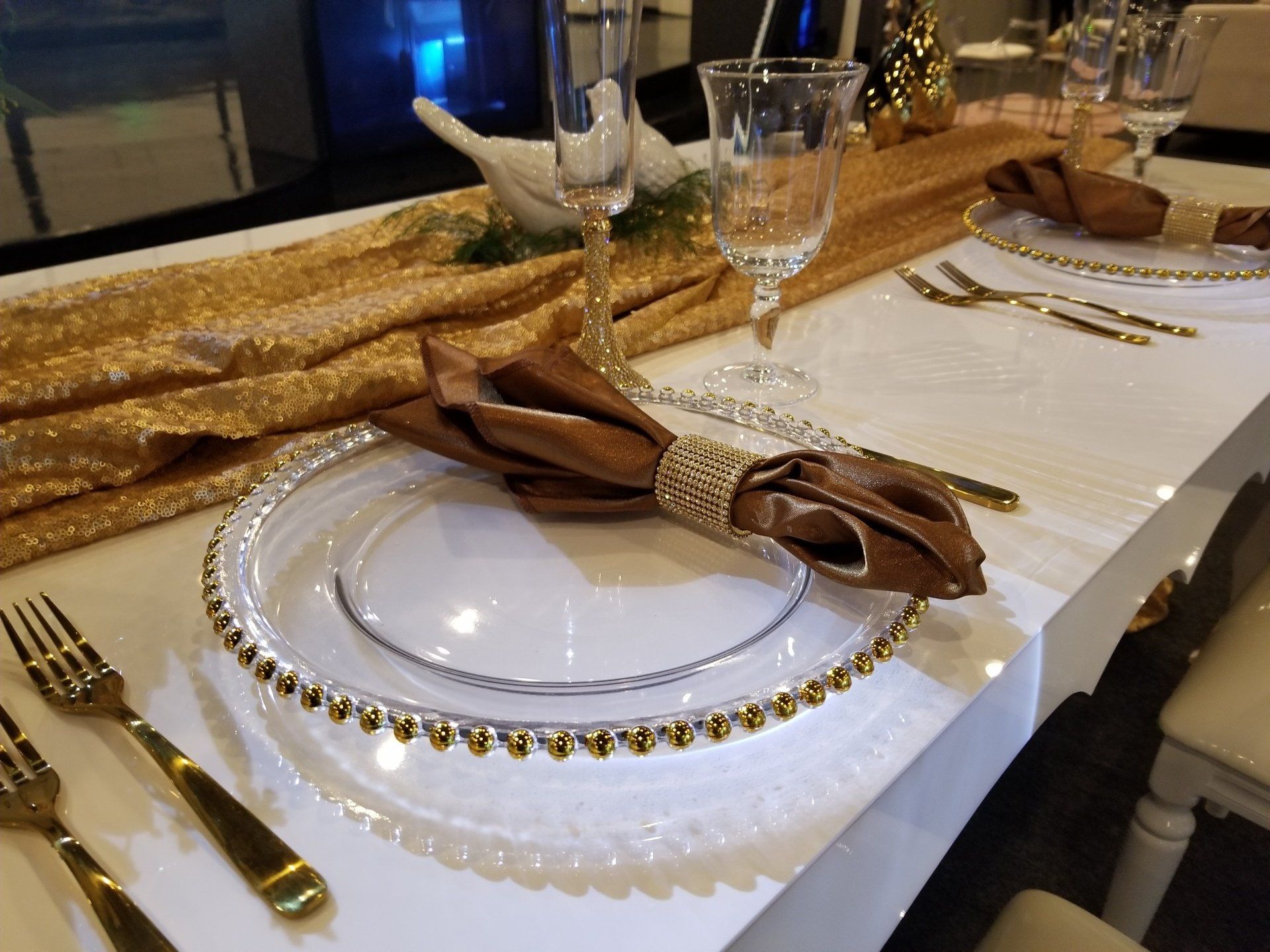 Formal table setting with gold accents, brown napkin, clear plate with gold trim, and gold silverware.
