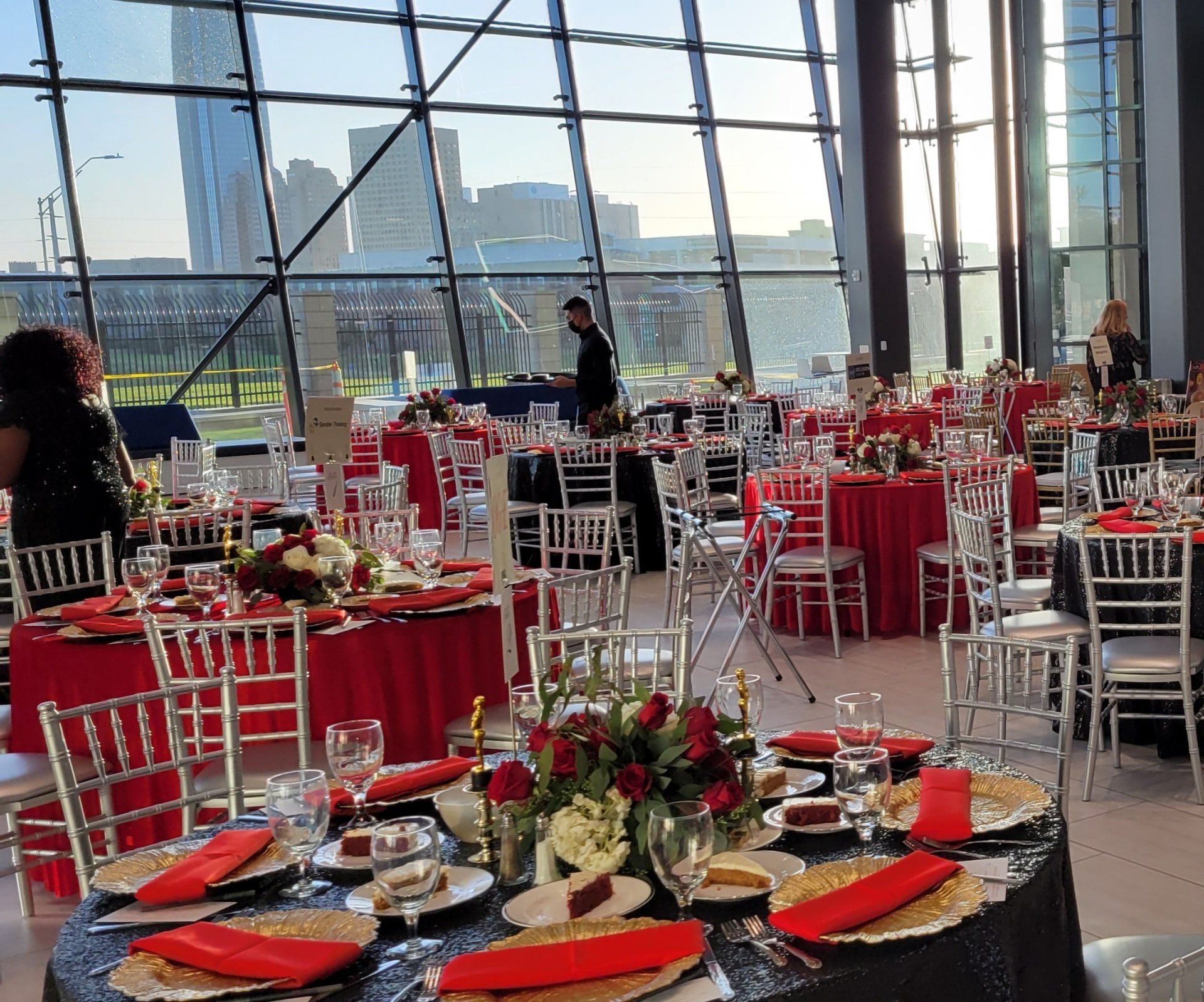 Tables set for an event with red and black linens, floral centerpieces, and silver chairs inside a modern building.