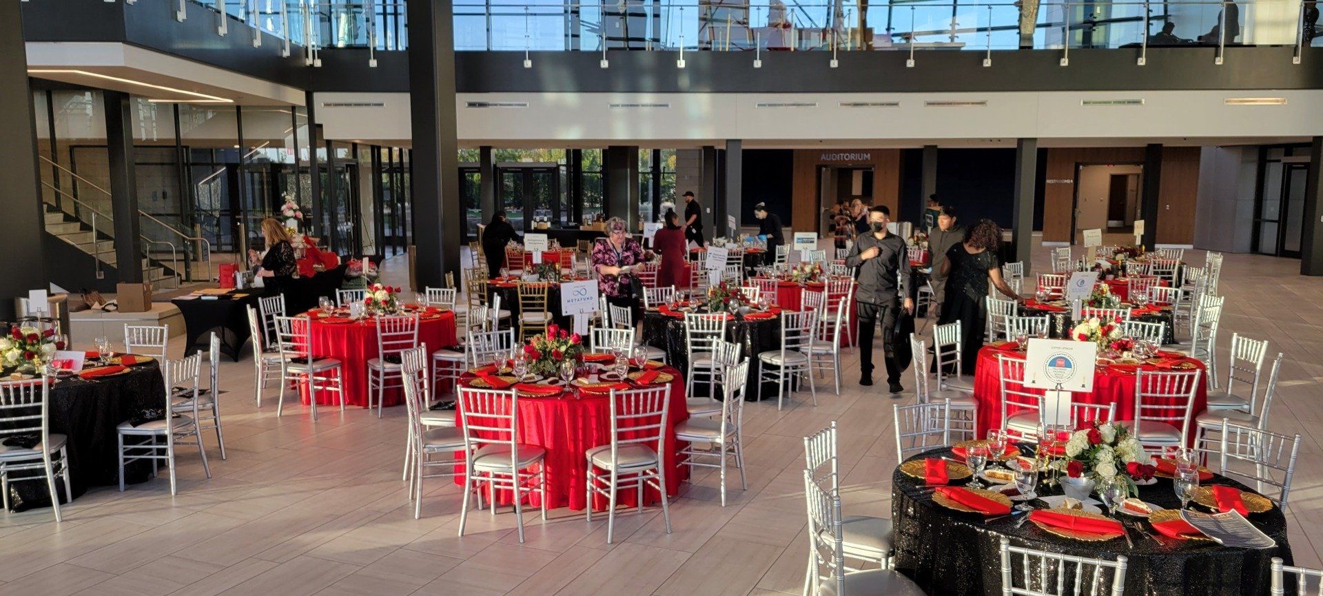 Tables with red and black tablecloths set for an event in a large hall with floor-to-ceiling windows.