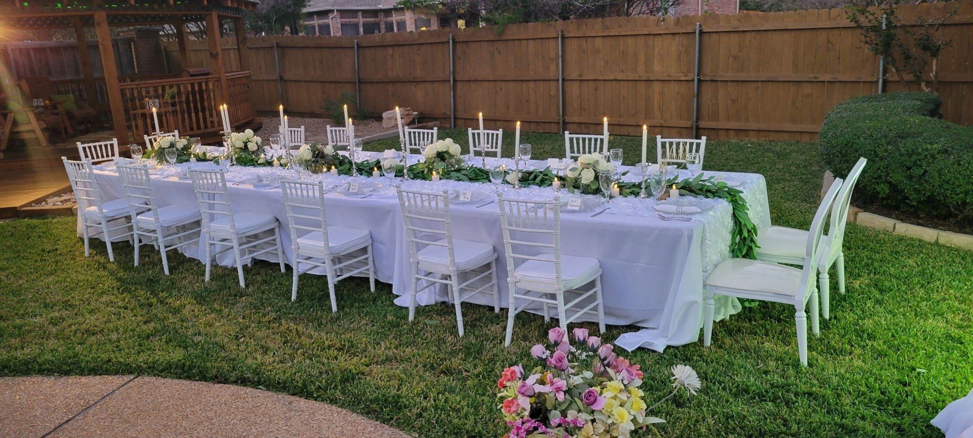 An outdoor dinner party setup with a long table, white chairs, and floral decorations on a lawn.